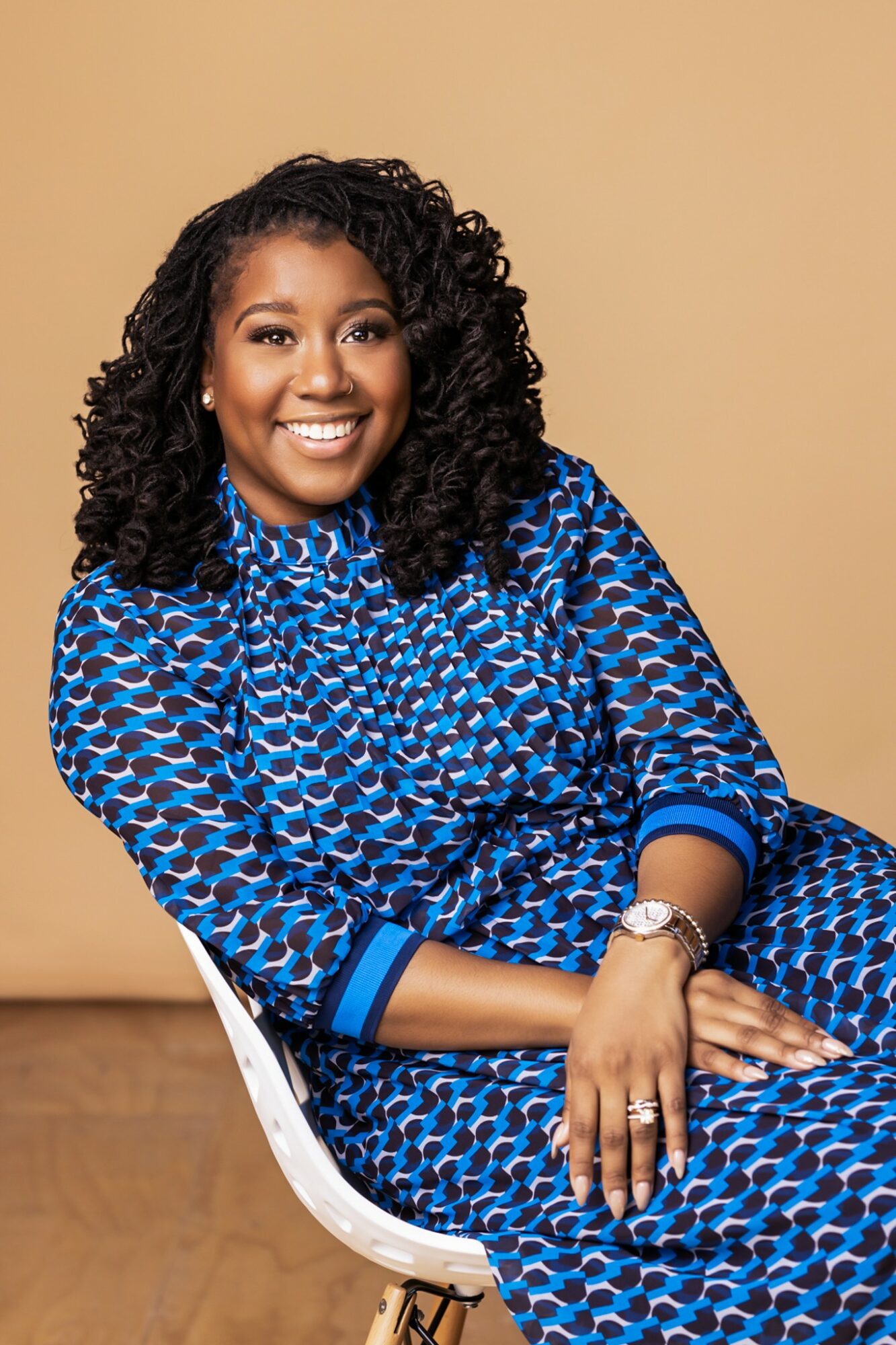 Smiling woman with curly hair wearing a blue patterned dress, sitting on a white chair, against a beige background.