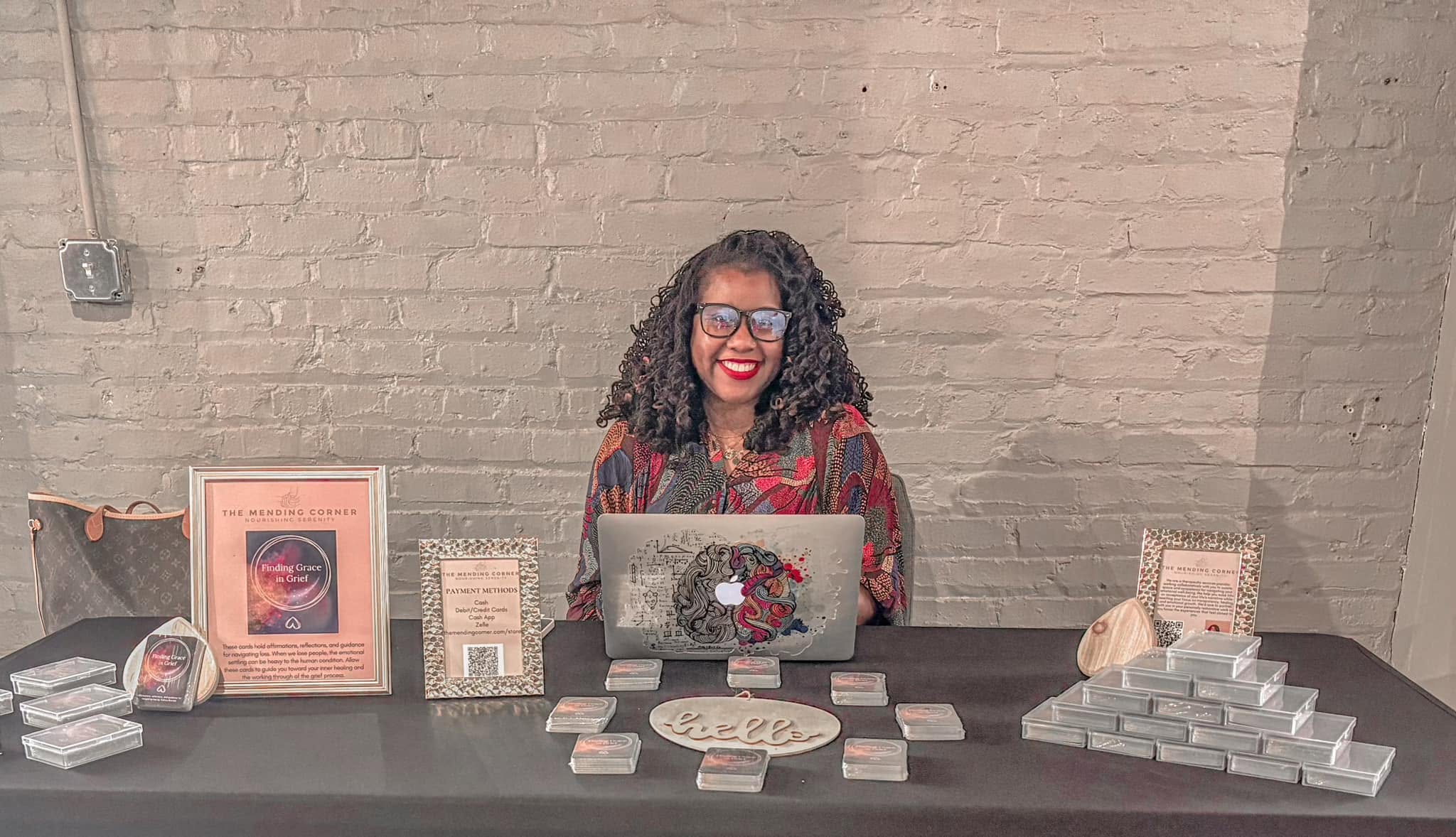 Woman with curly hair and glasses sitting at a table with cards and signs, smiling, against a brick wall.