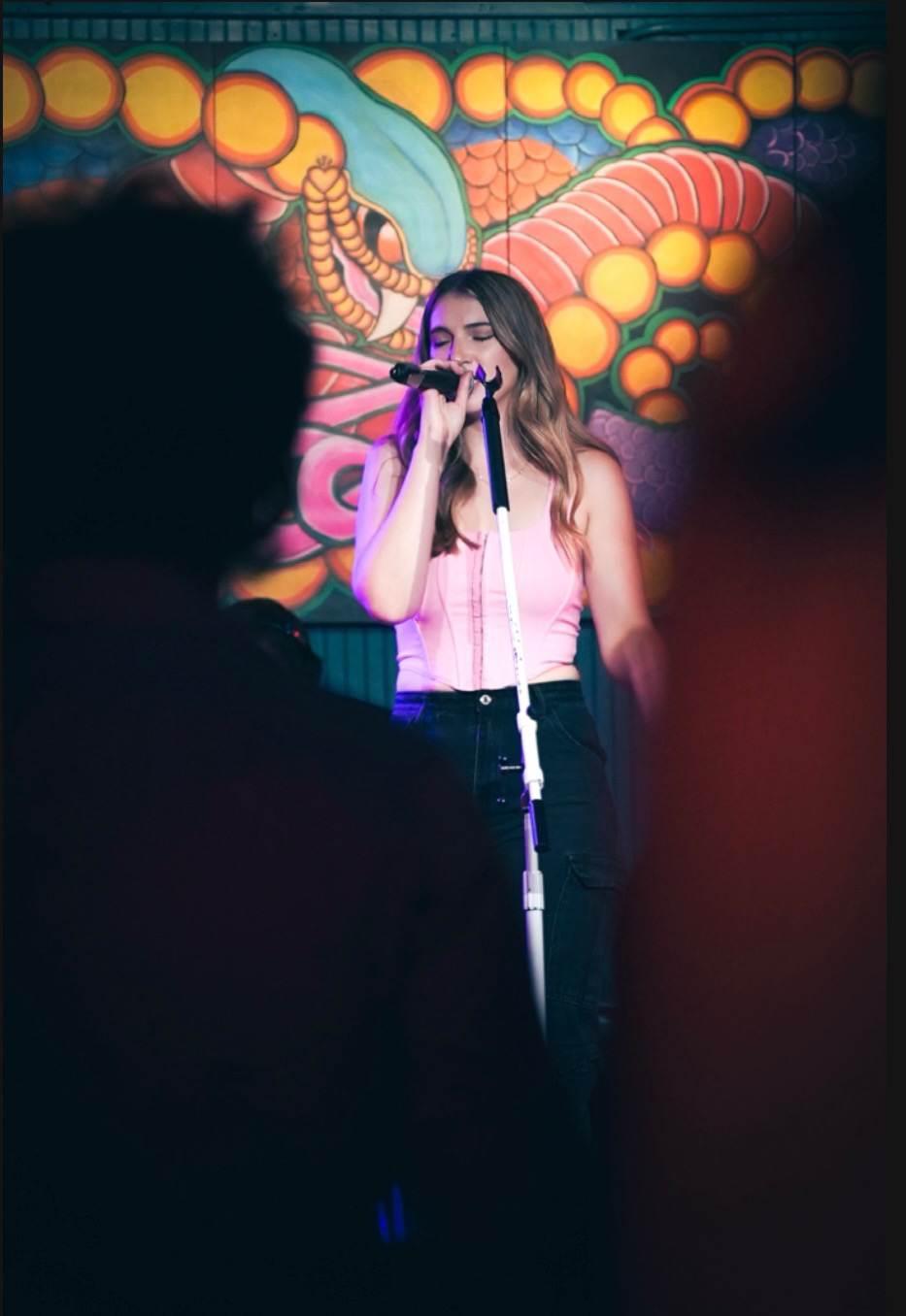 A woman singing into a microphone on stage with colorful abstract background, audience in foreground, vibrant atmosphere.