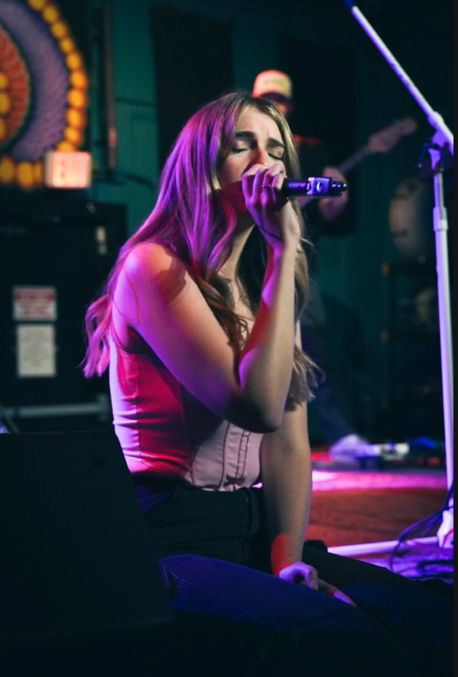 A woman with long hair sings into a microphone on stage, with colorful lighting and equipment in the background.