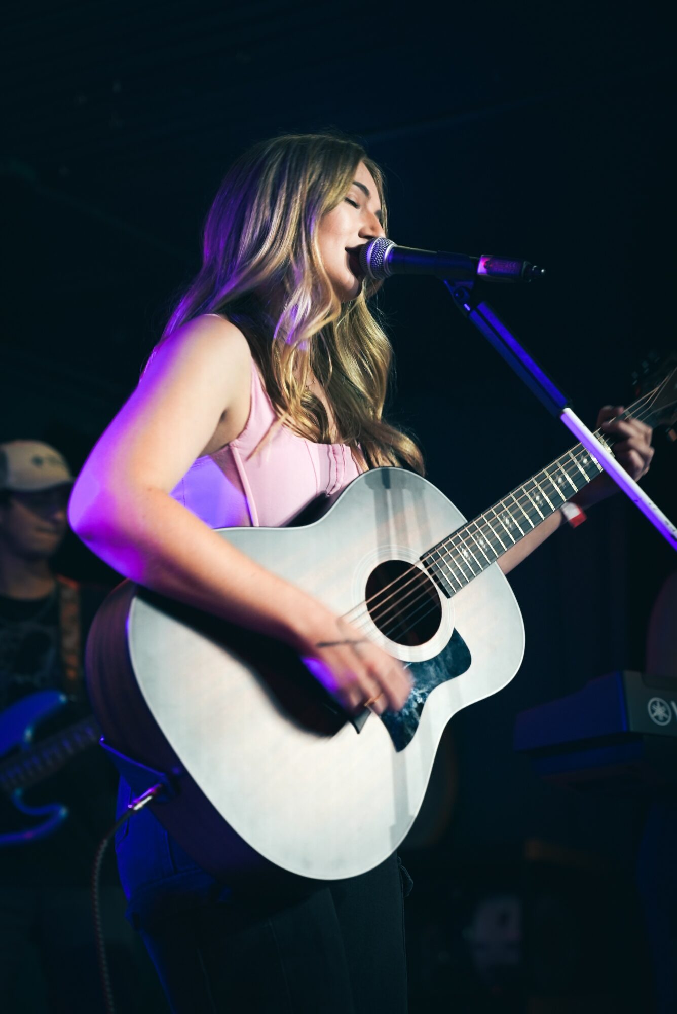 Woman with long hair singing into microphone while playing acoustic guitar on stage.