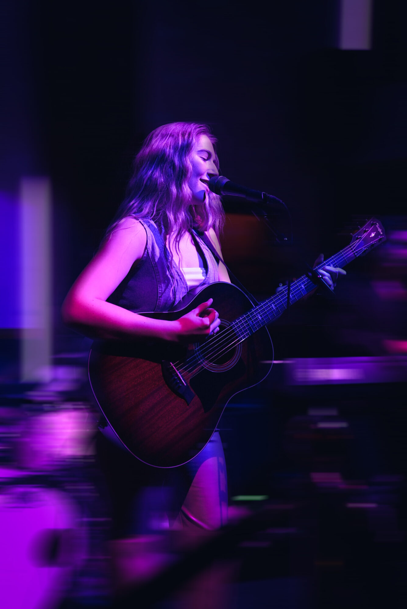 Young woman playing guitar and singing into microphone on stage with purple lighting, dark background, and blurred audience.