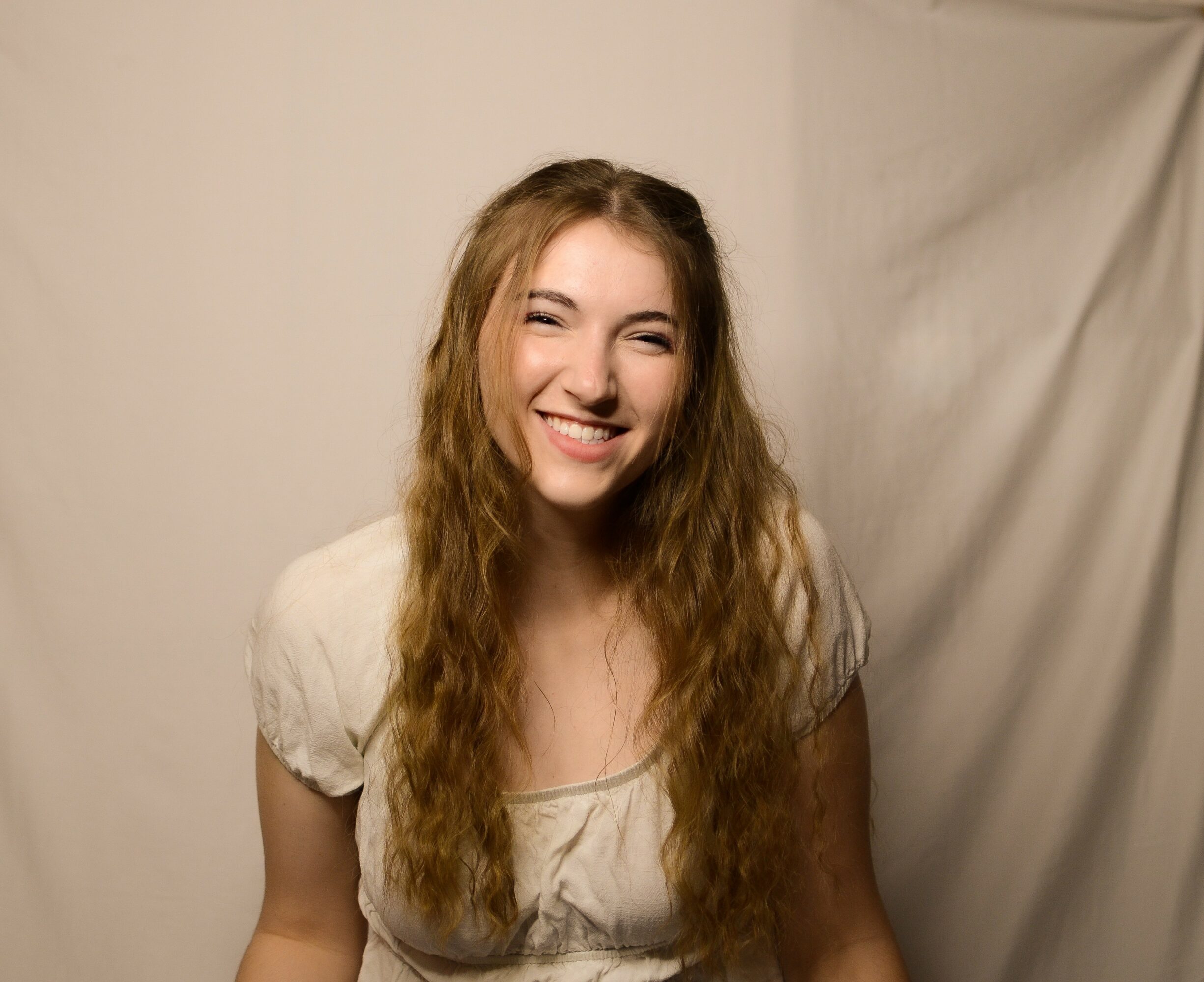 Young woman with long wavy hair smiling, wearing a light-colored top, standing against a plain background.