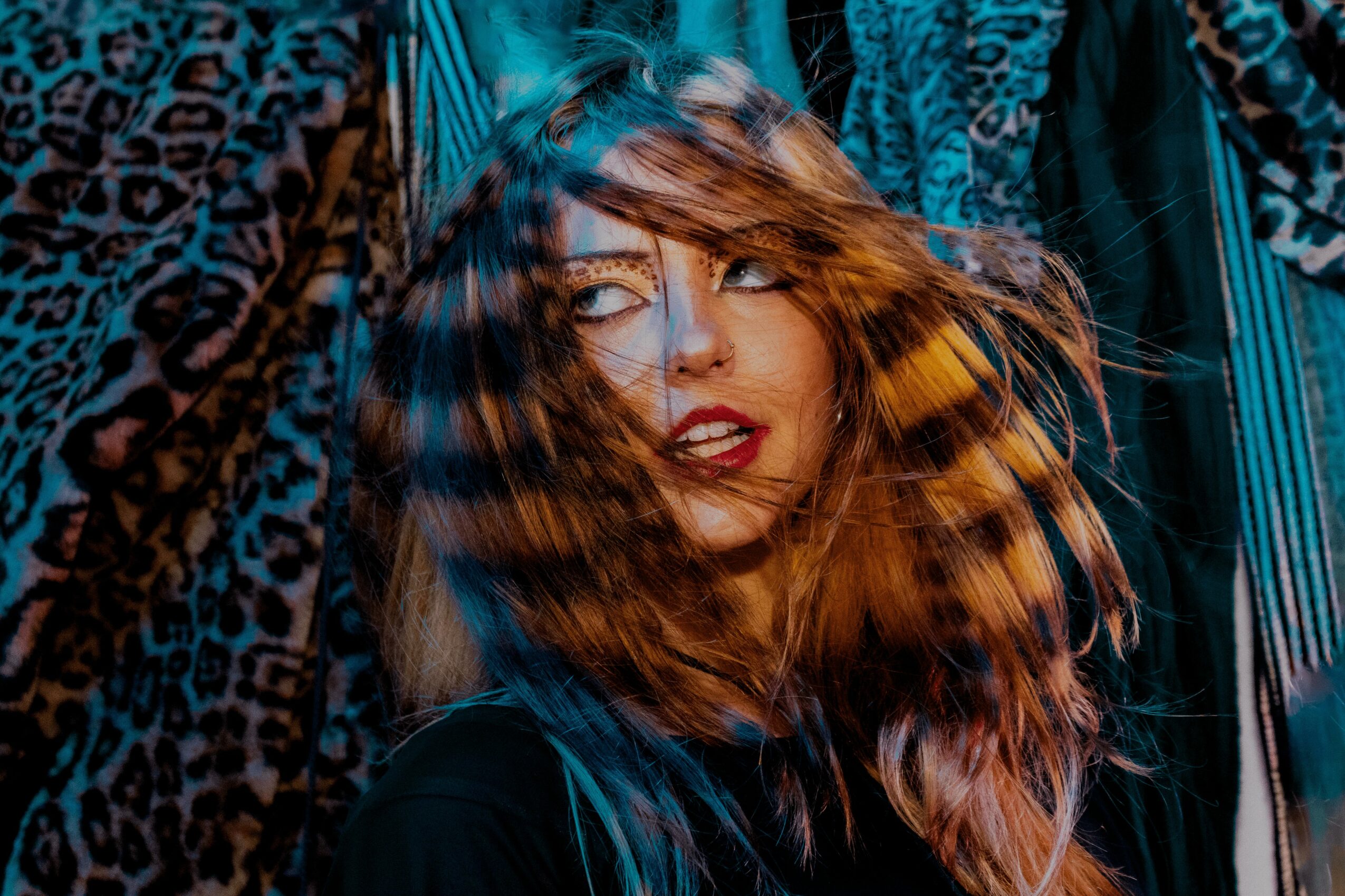 Woman with long, wavy hair and makeup, surrounded by colorful fabrics or clothing, looking to the side.
