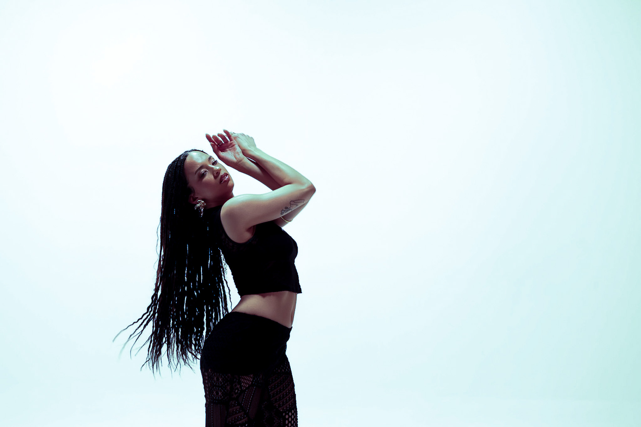 Woman with long hair in black outfit holding a phone to her ear, standing sideways against a light background.
