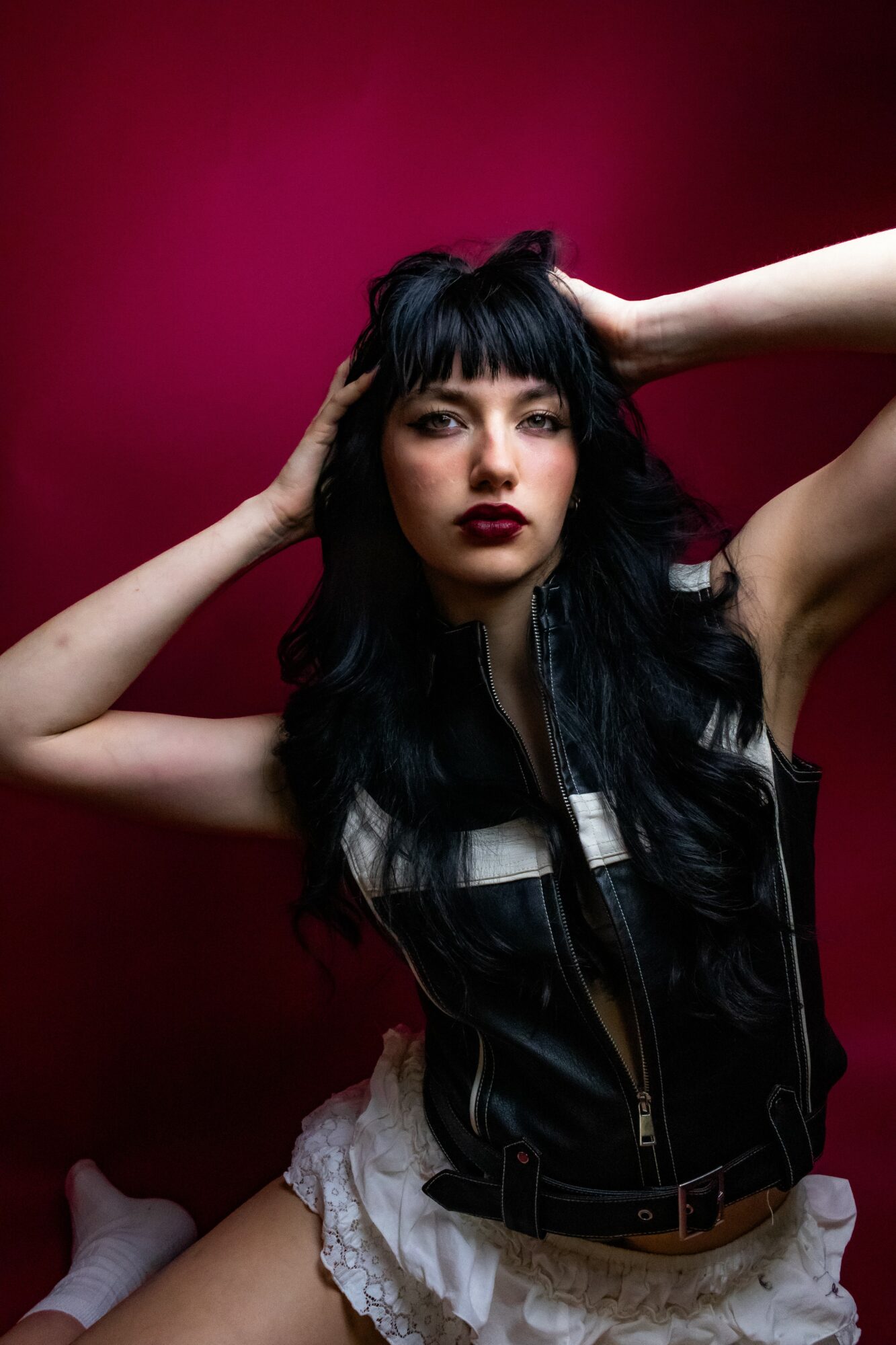 Woman with long black hair and dark lipstick poses with hands on head against a dark pink background.