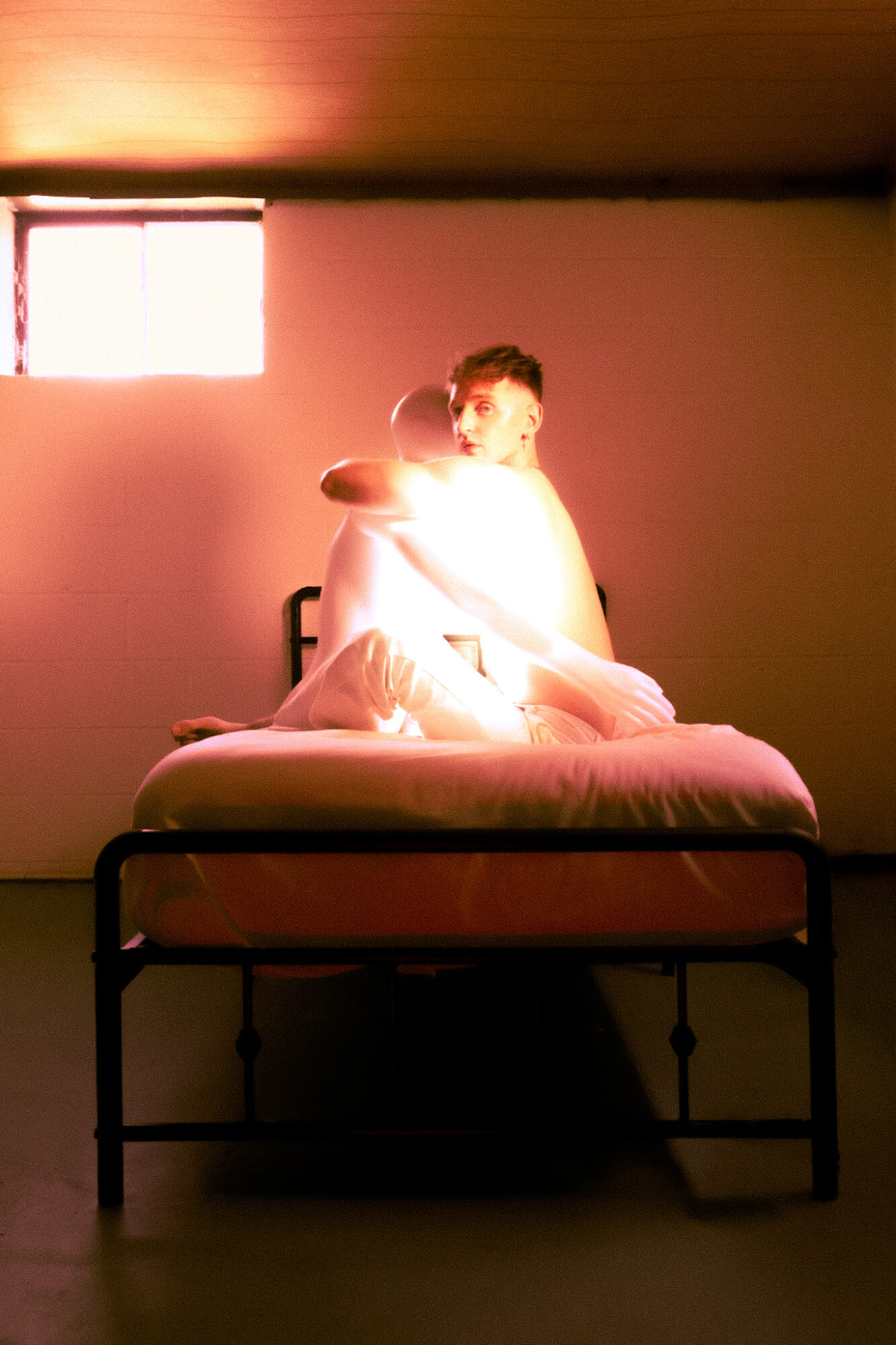 Person sitting on a bed in a dimly lit room with a small window, facing the camera, illuminated by warm light.