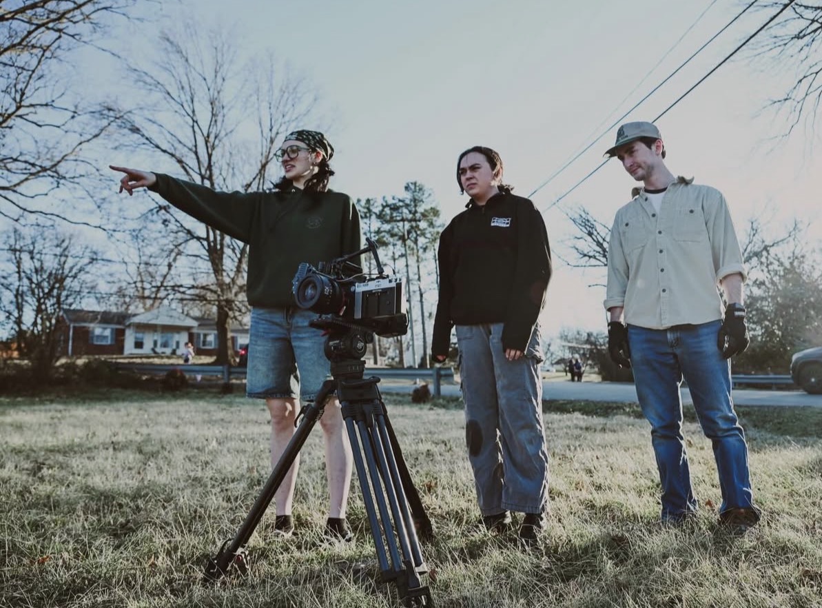 Three people outdoors, one pointing, with a camera on a tripod, trees, and houses in the background.