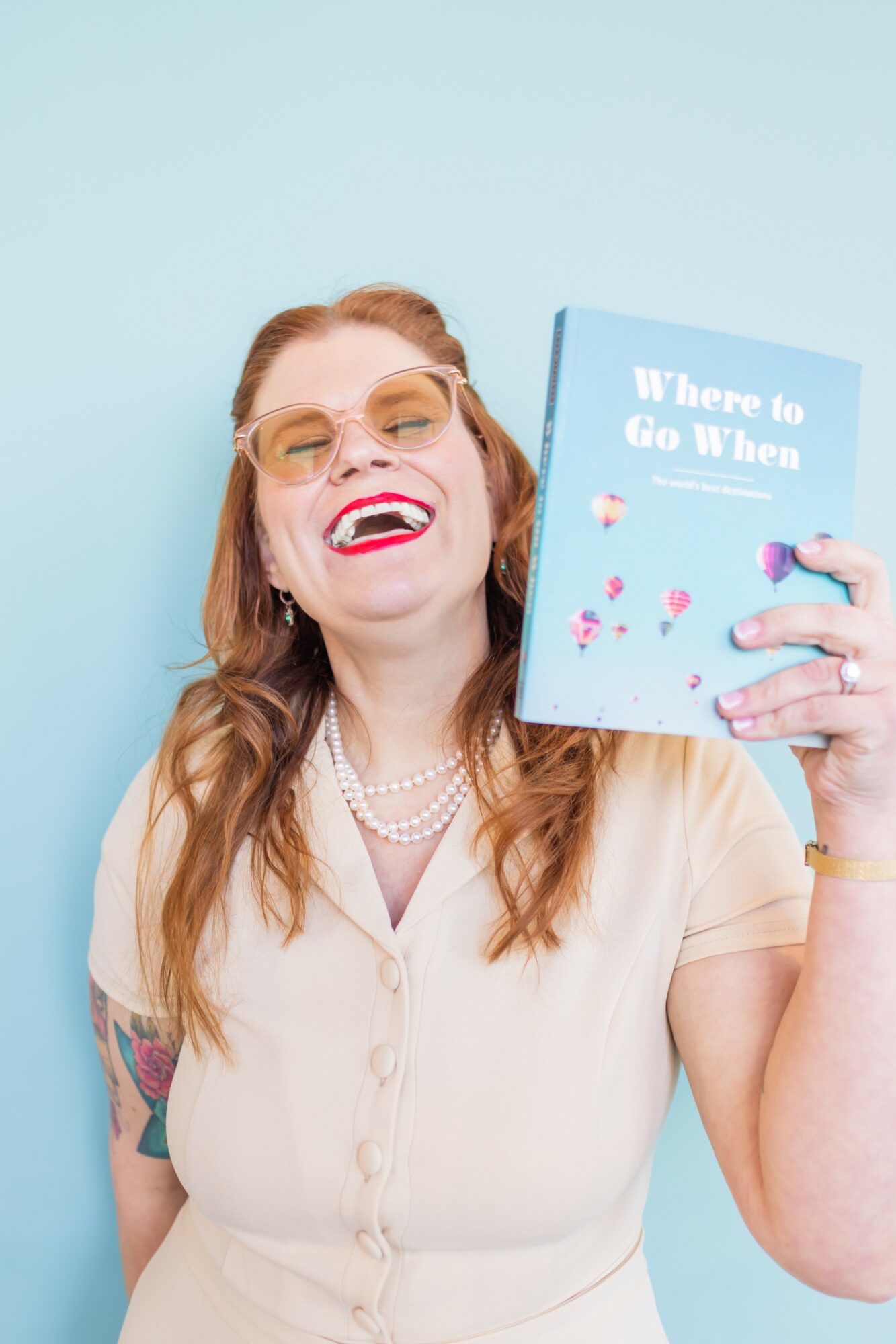 Woman with glasses and pearl necklace smiling, holding a book titled 'Where to Go When' against a light blue background.