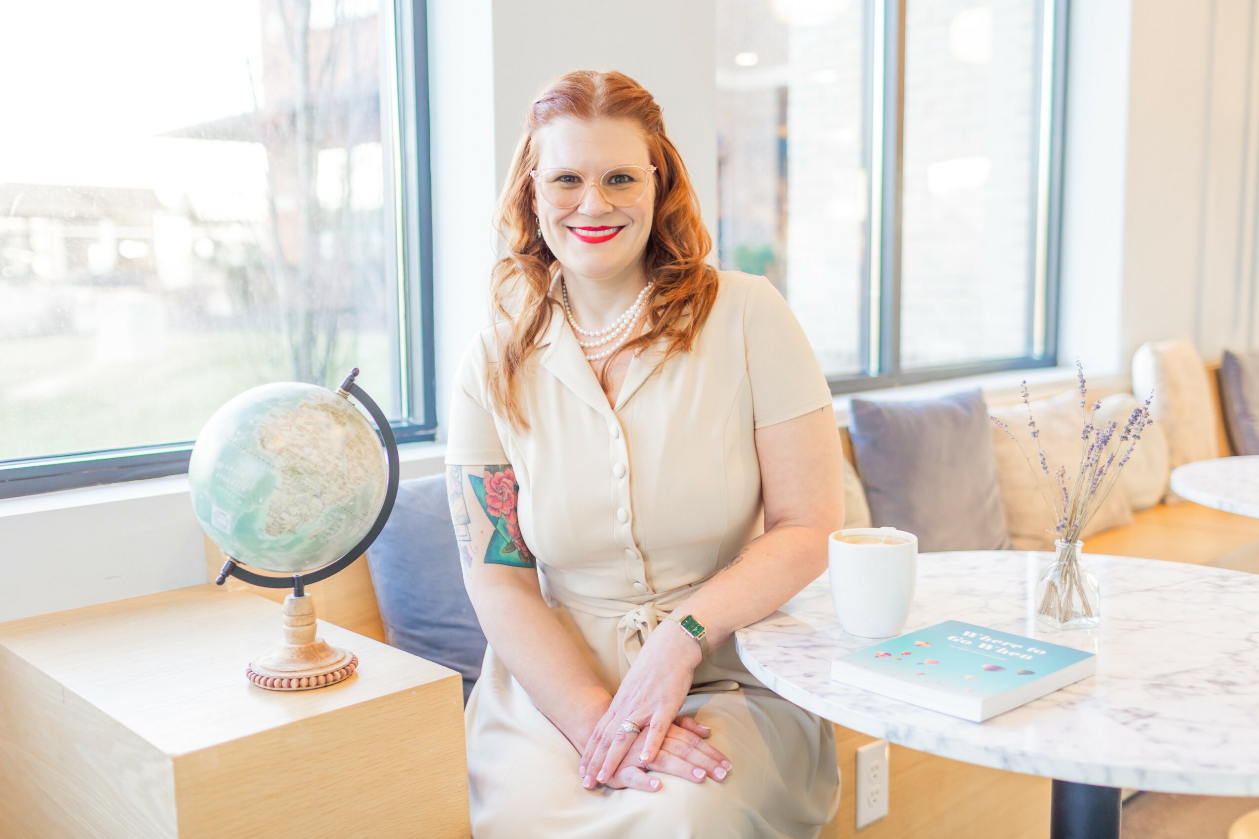 Woman with red hair and a tattoo on her arm, smiling, sitting at a table near a window, with a globe and a book nearby.