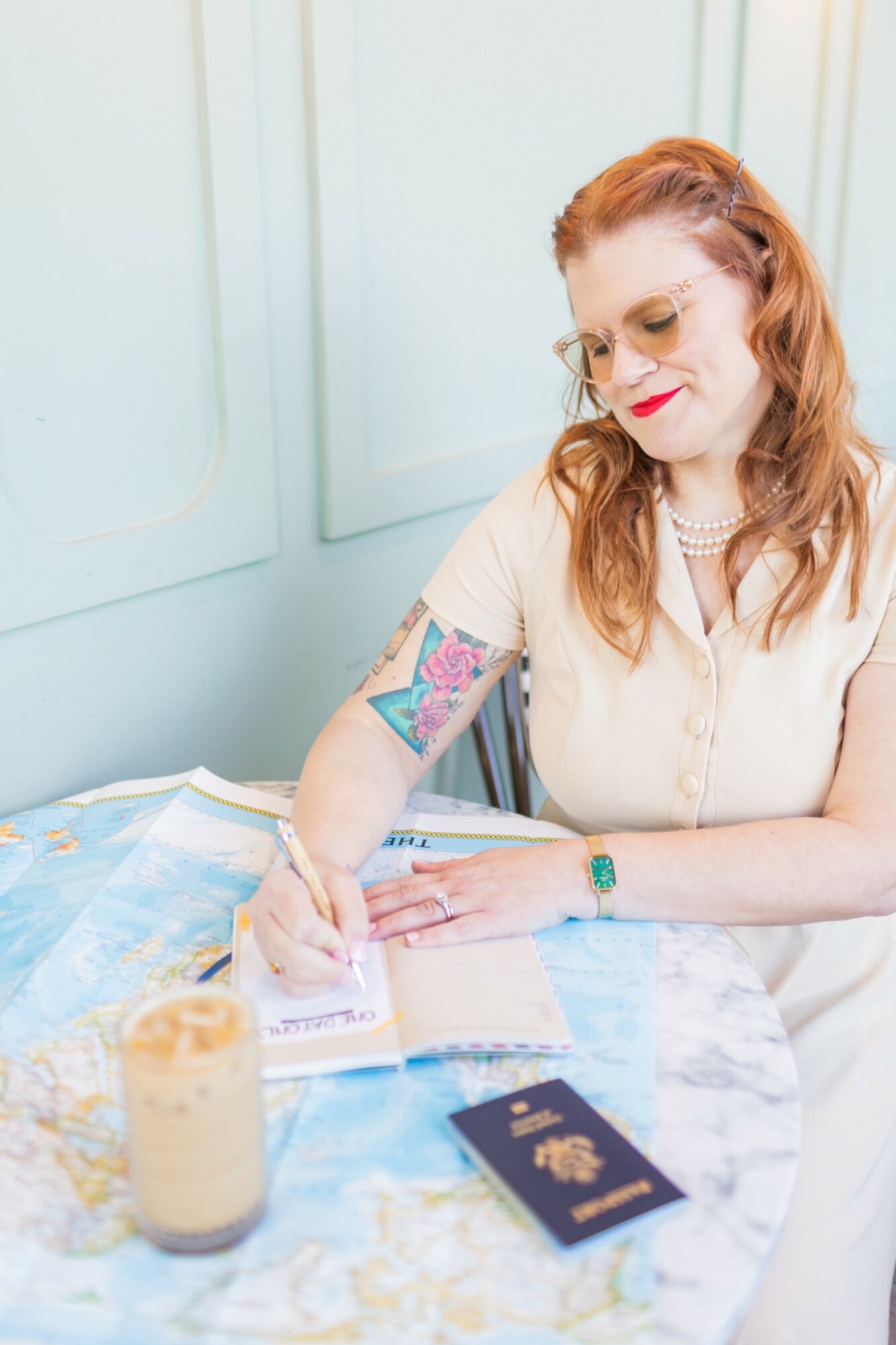 Woman with red hair and glasses writing in a notebook at a table with a map, a drink, and a passport.