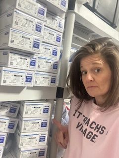 Woman with shoulder-length hair wearing a pink shirt with text, standing next to stacked boxes in a store.
