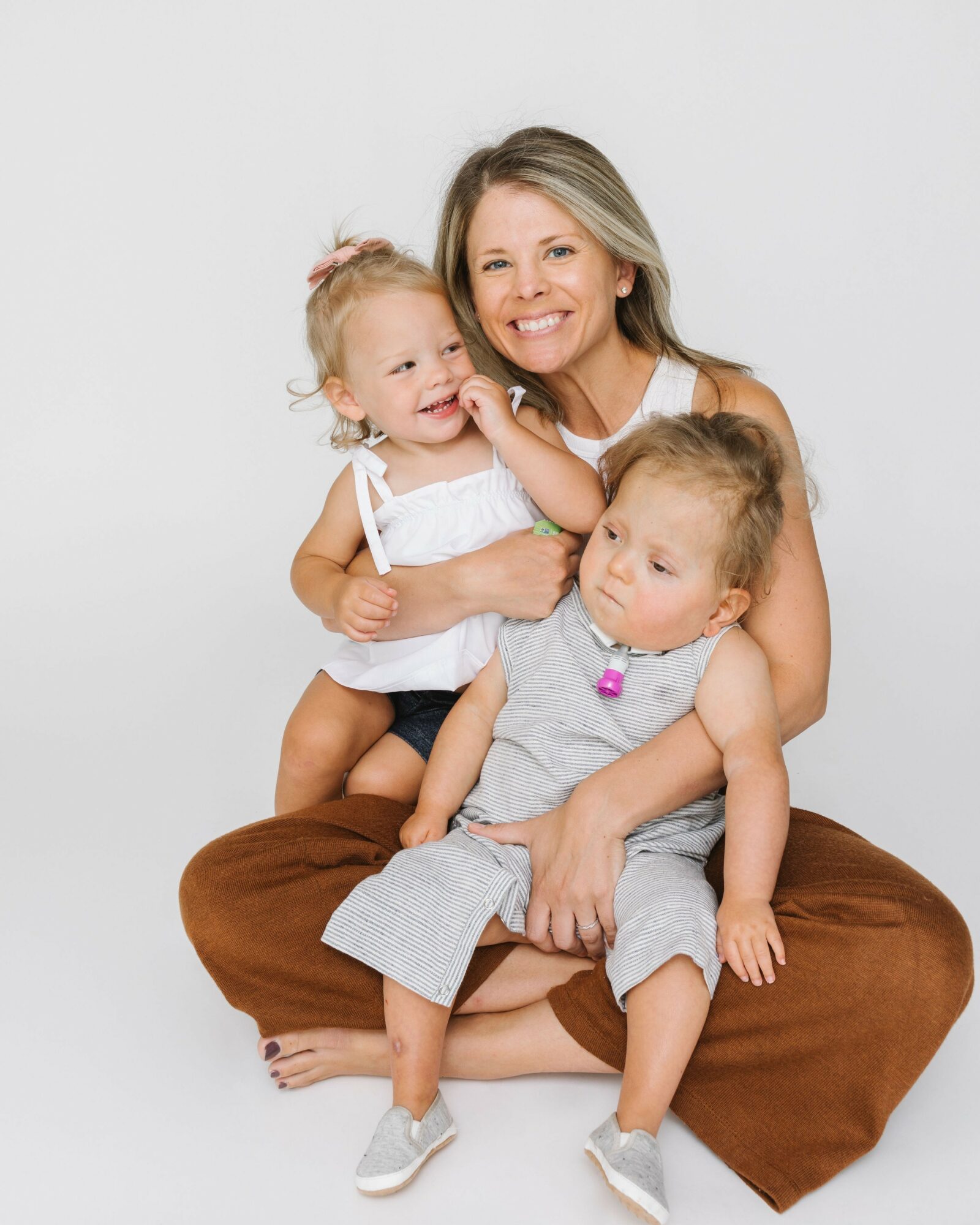 Woman with two young children sitting on a cushion, smiling and playing together, against a plain white background.
