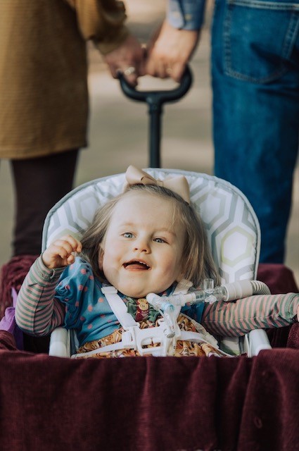 Young girl in a stroller smiling, holding hands with two adults, outdoors, with a blurred background.