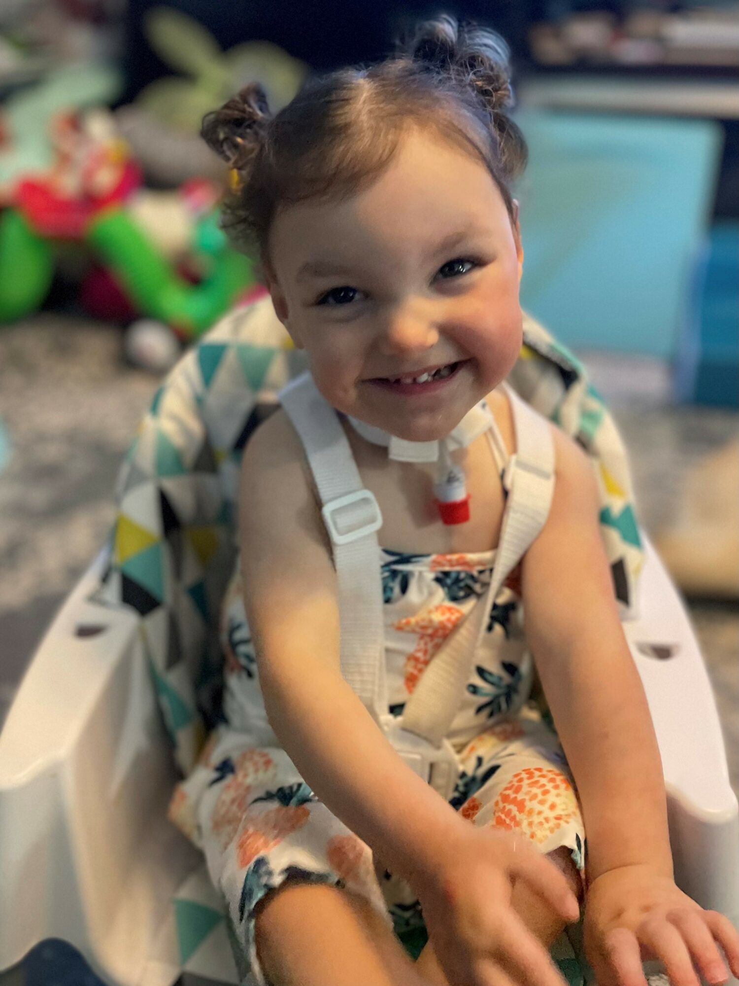 Smiling young girl with curly hair in a high chair, wearing a colorful dress and a medical device around her neck.