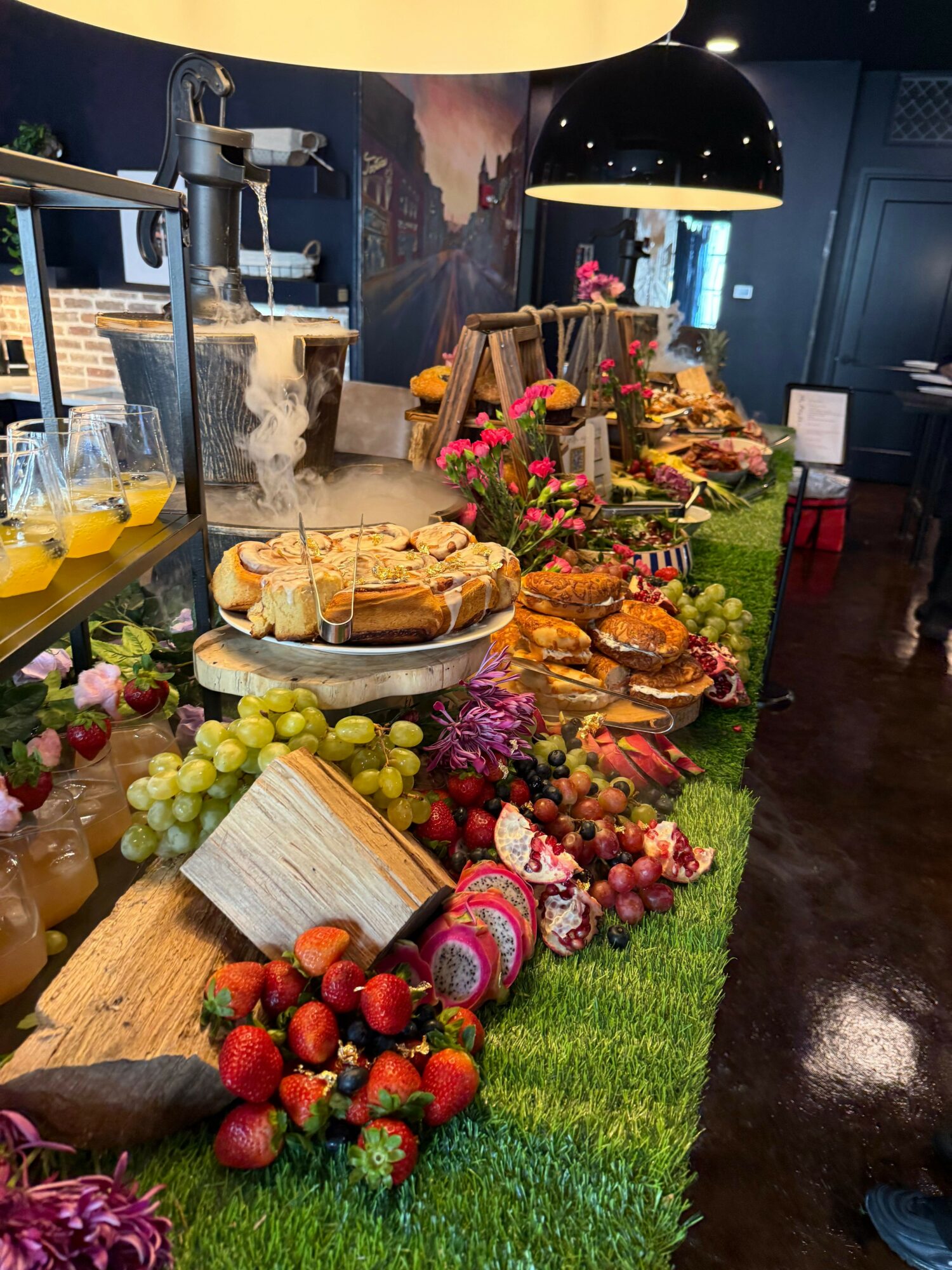 Display of fruits, bread, and flowers on a table with green grass-like surface, arranged decoratively.
