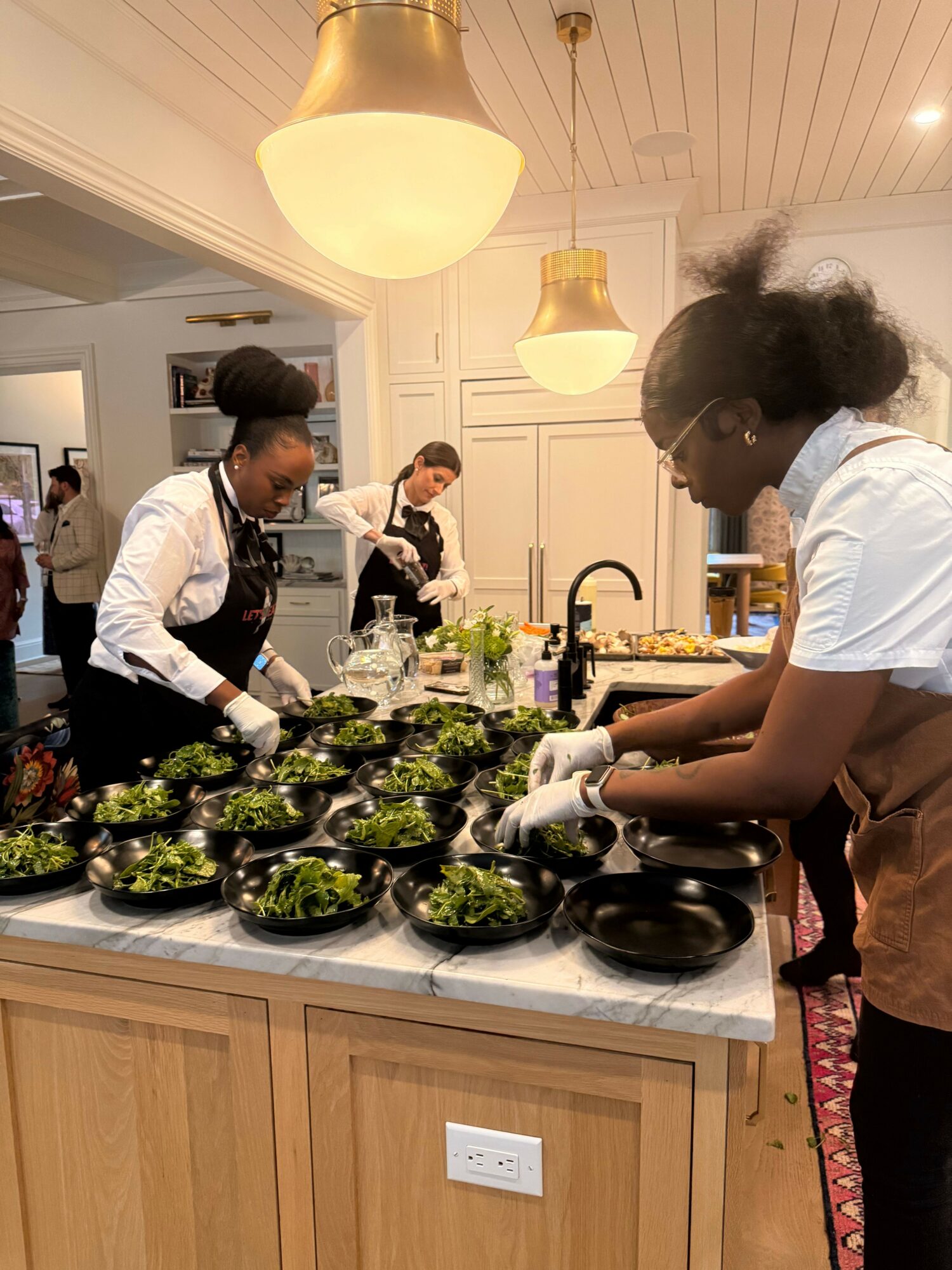 People preparing food in a kitchen with a woman washing vegetables and a man in a vest and tie at the counter, with a third person in the background. The kitchen has white cabinets and a wooden countertop.