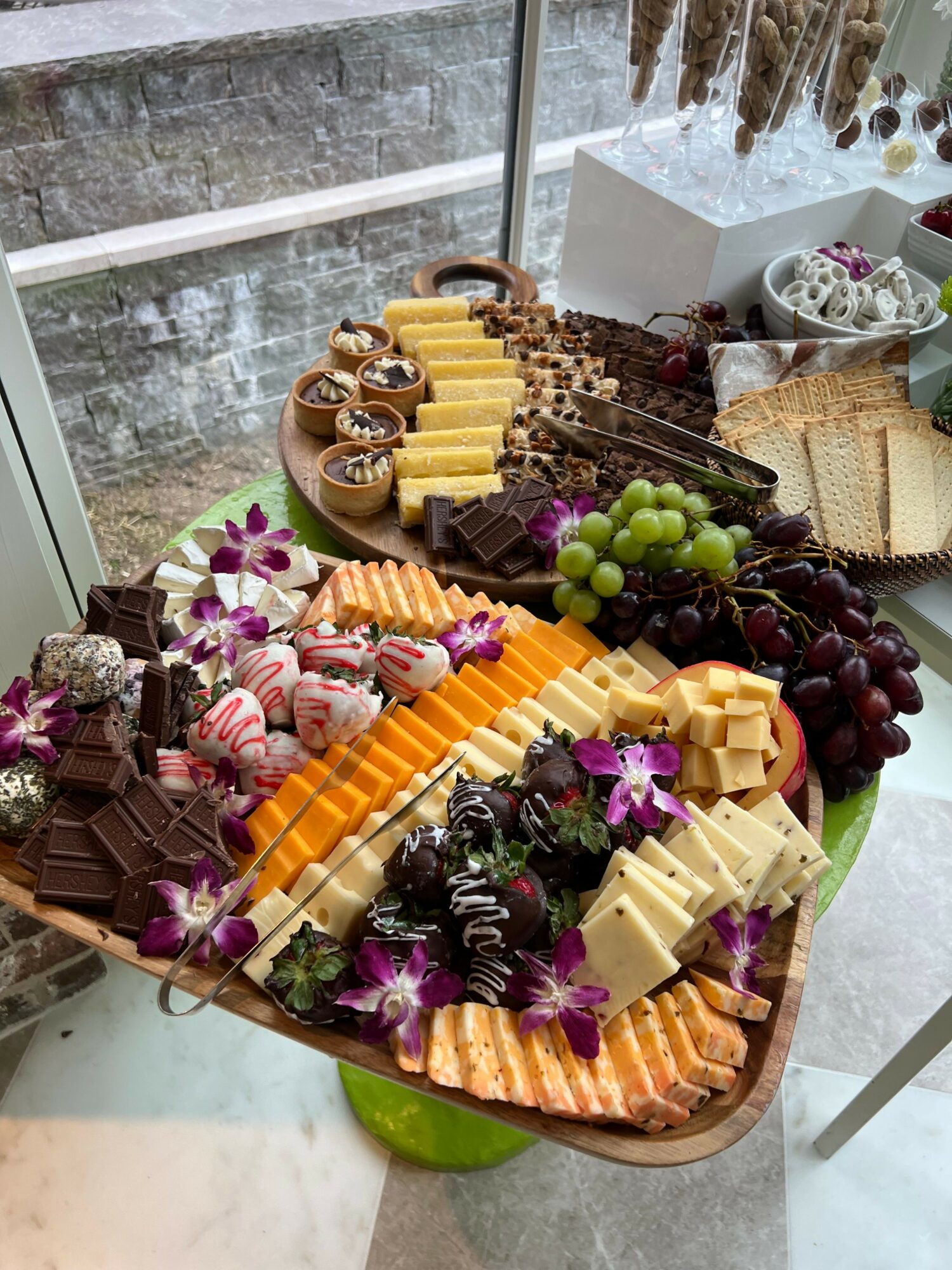 Cheese, grapes, and chocolates arranged on a wooden tray with purple flowers, on a table near a window.
