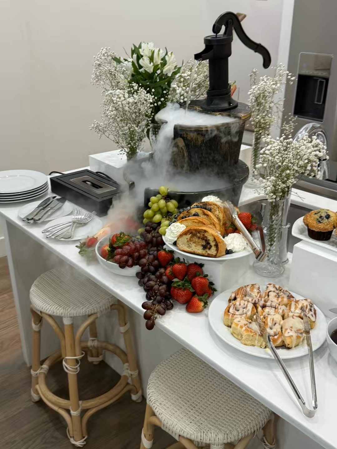 Table with flowers, baked goods, grapes, and a decorative water fountain, with chairs nearby.