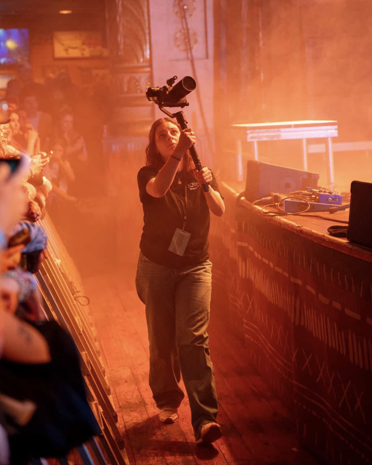 A woman holding a camera on a tripod in a smoky, orange-lit indoor event with a crowd on the left side.
