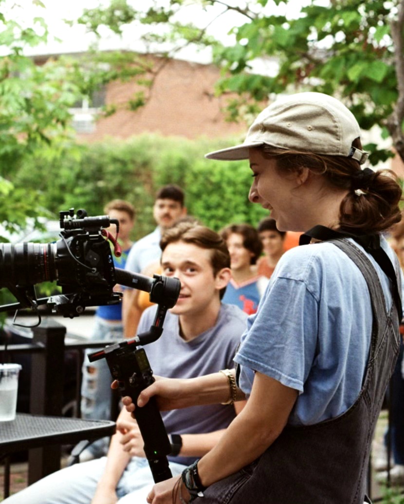 Person operating camera equipment outdoors with group of people in background, surrounded by trees and greenery.