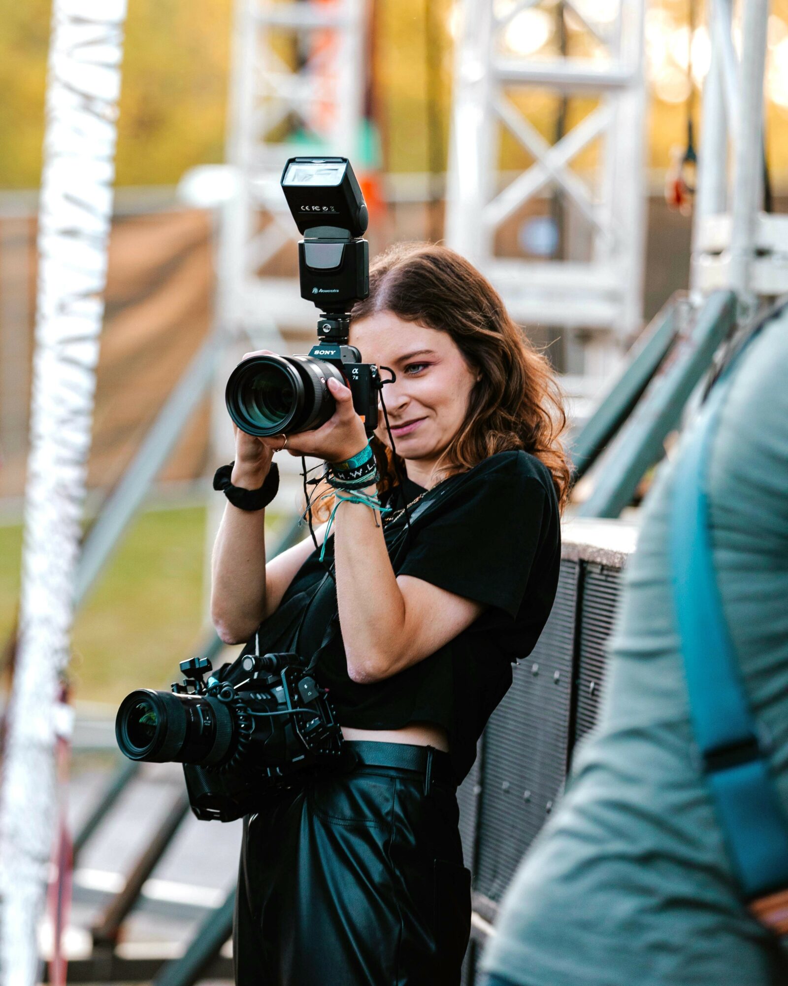 Young woman holding a camera with a flash, smiling, outdoors with trees and metal structures in background.