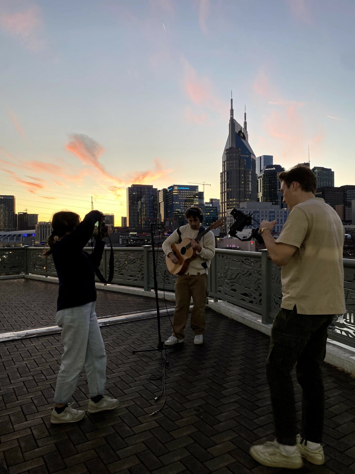 Four people on a rooftop with a city skyline and sunset in the background, one playing guitar, others taking photos.