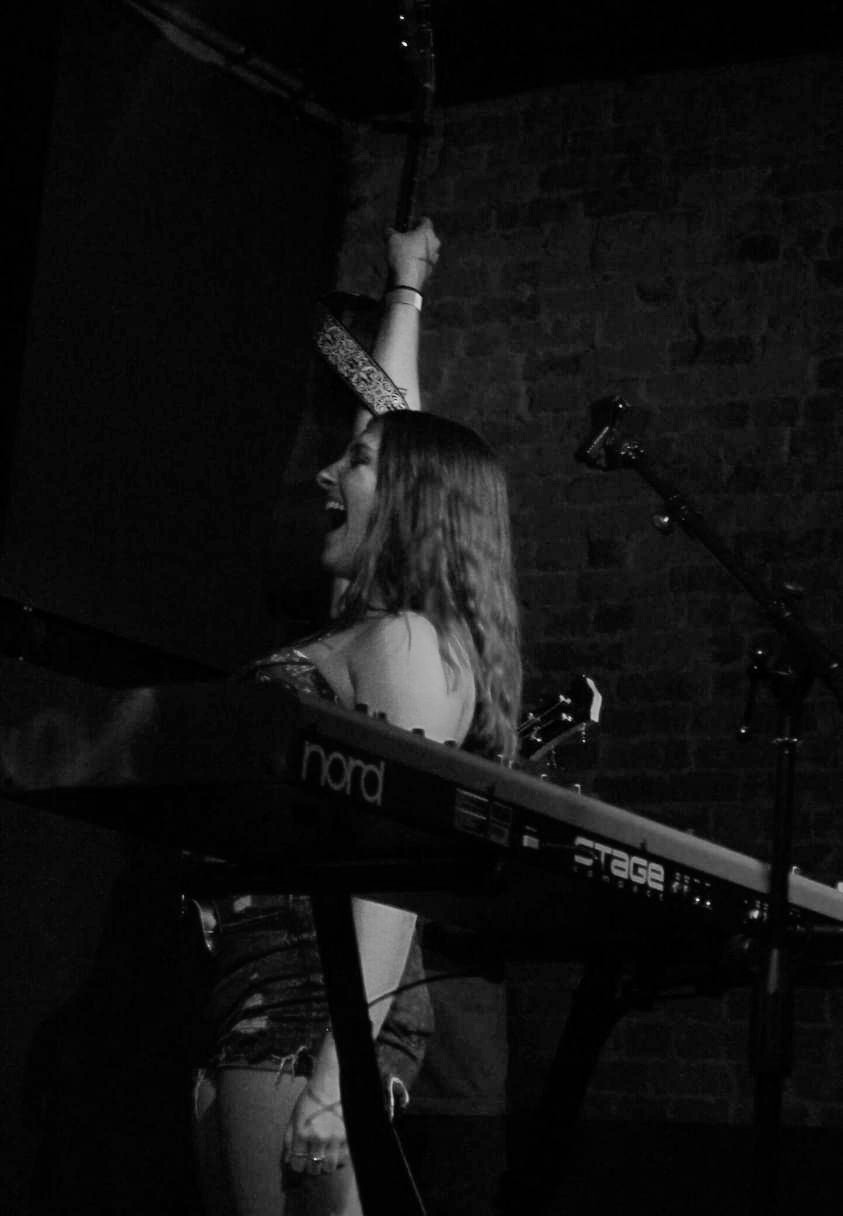 Person with long hair raising an arm, standing behind a keyboard on a stage with a brick wall background.