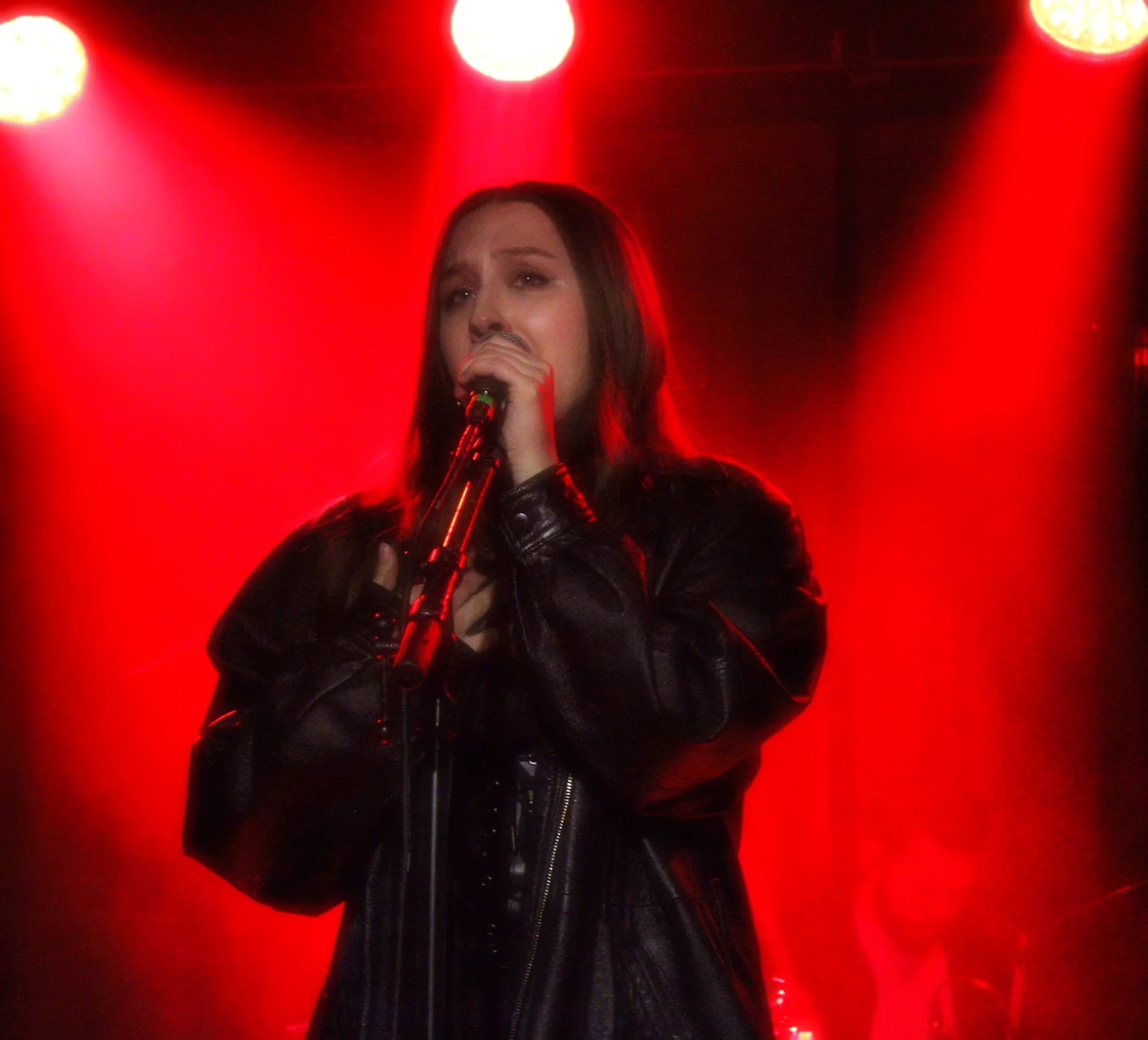 A woman singing into a microphone on stage with red lighting behind her.