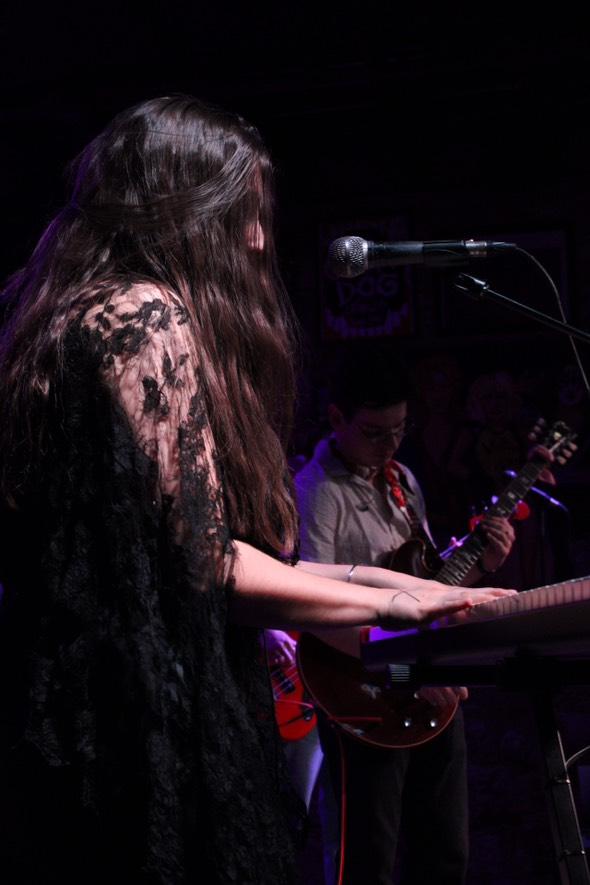 A woman with long hair wearing a lace dress plays a keyboard on stage, with a guitarist in the background.