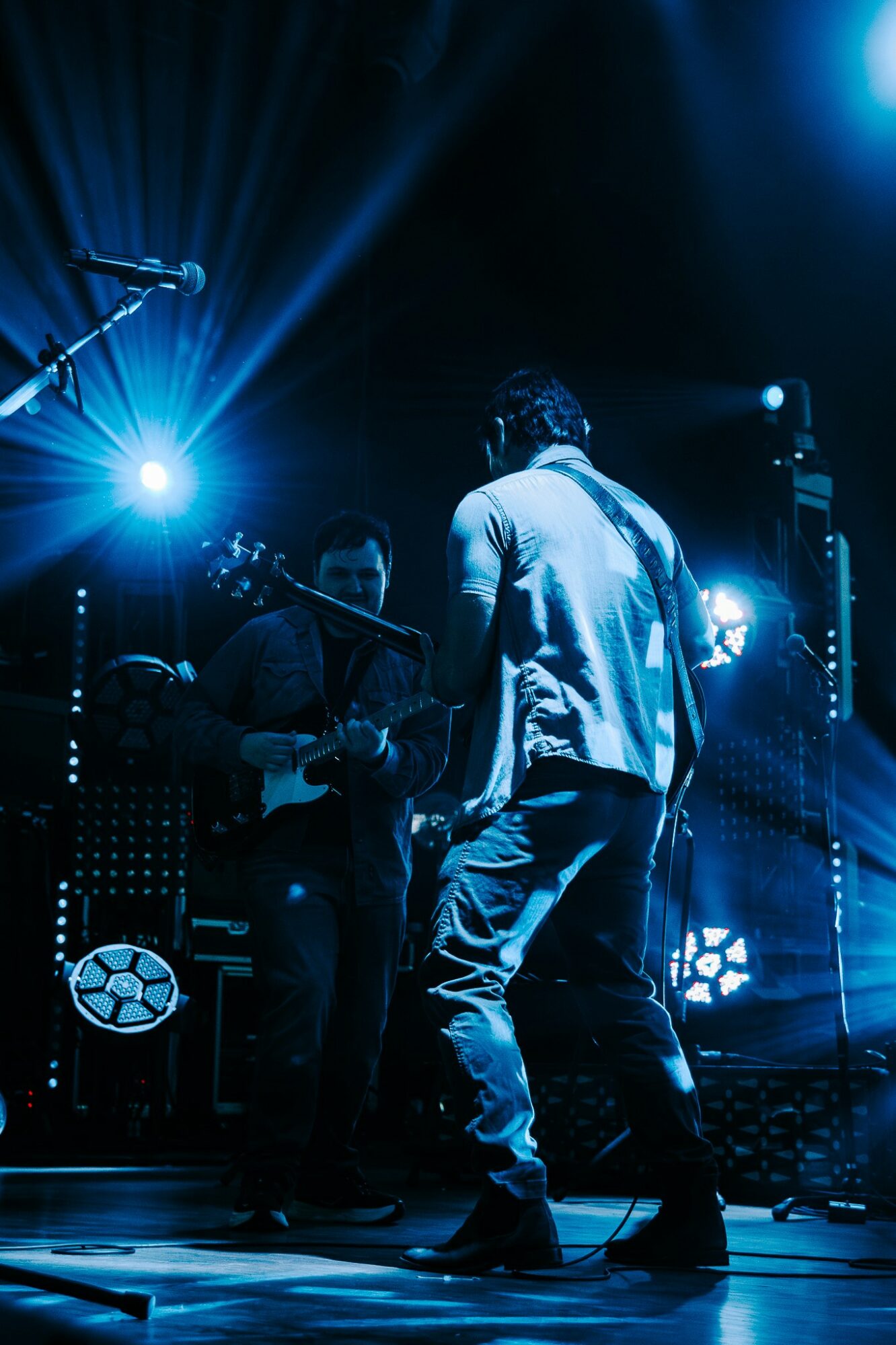 Two musicians perform on stage with bright blue and white lights, one playing guitar and the other singing or speaking.