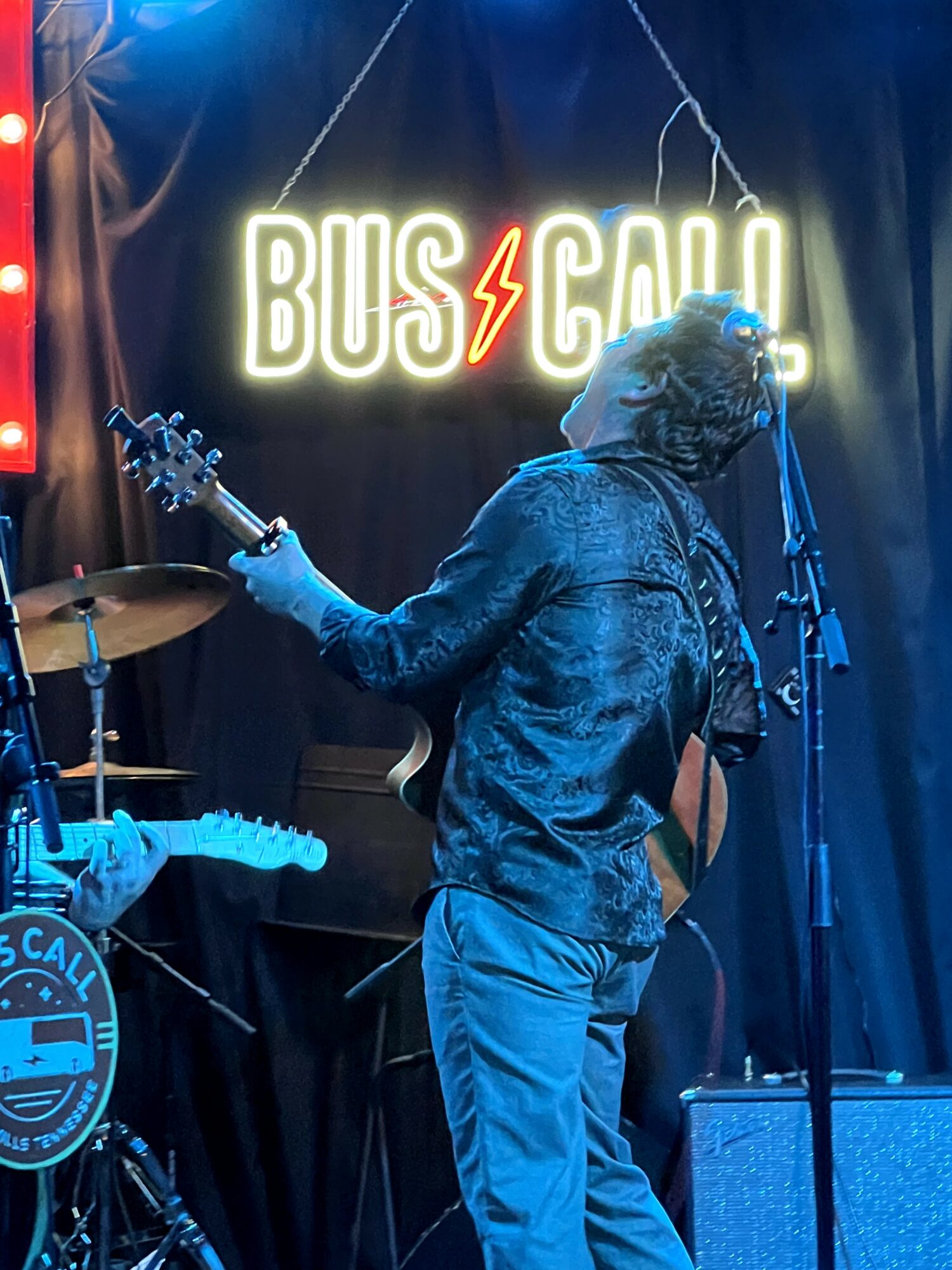 Person playing guitar on stage with a neon sign reading 'BUS⚡SCAL' in the background.
