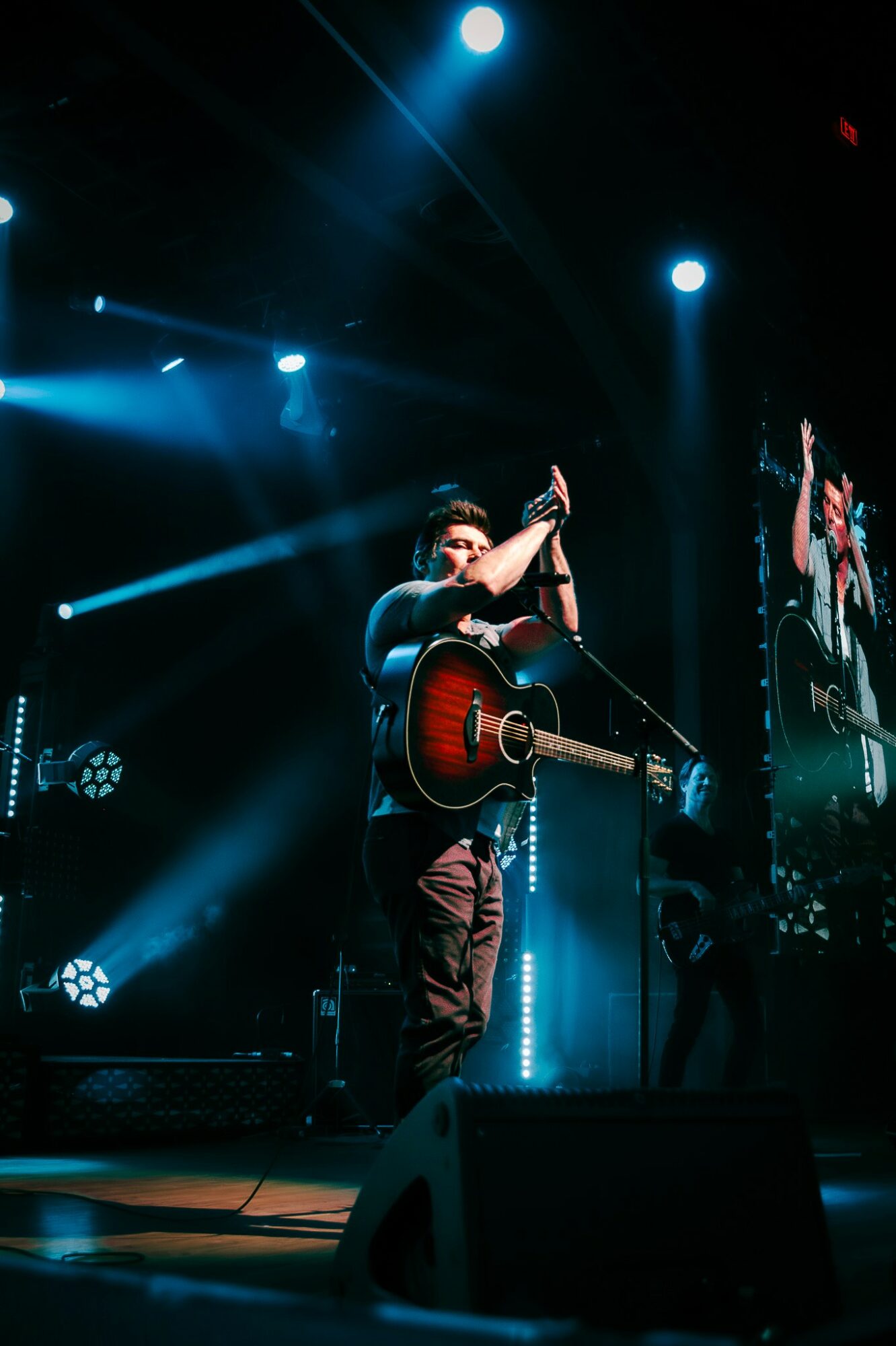 Performer on stage with guitar, clapping, illuminated by blue stage lights, large screen in background, dark setting.