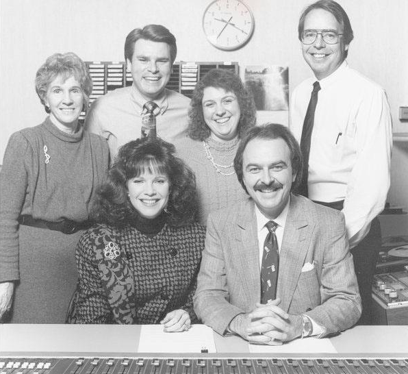 Group of seven people in an office, smiling, with a clock on the wall behind them.