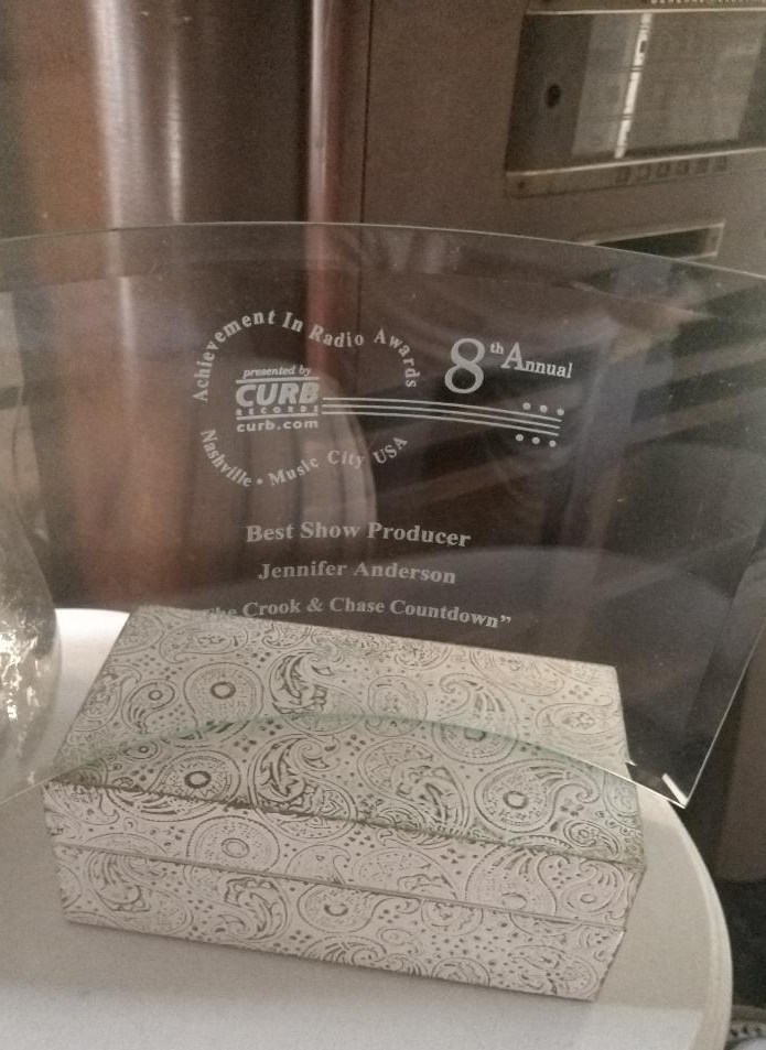 Clear glass award with engraved text, placed on a patterned box, background includes metallic and glass surfaces.