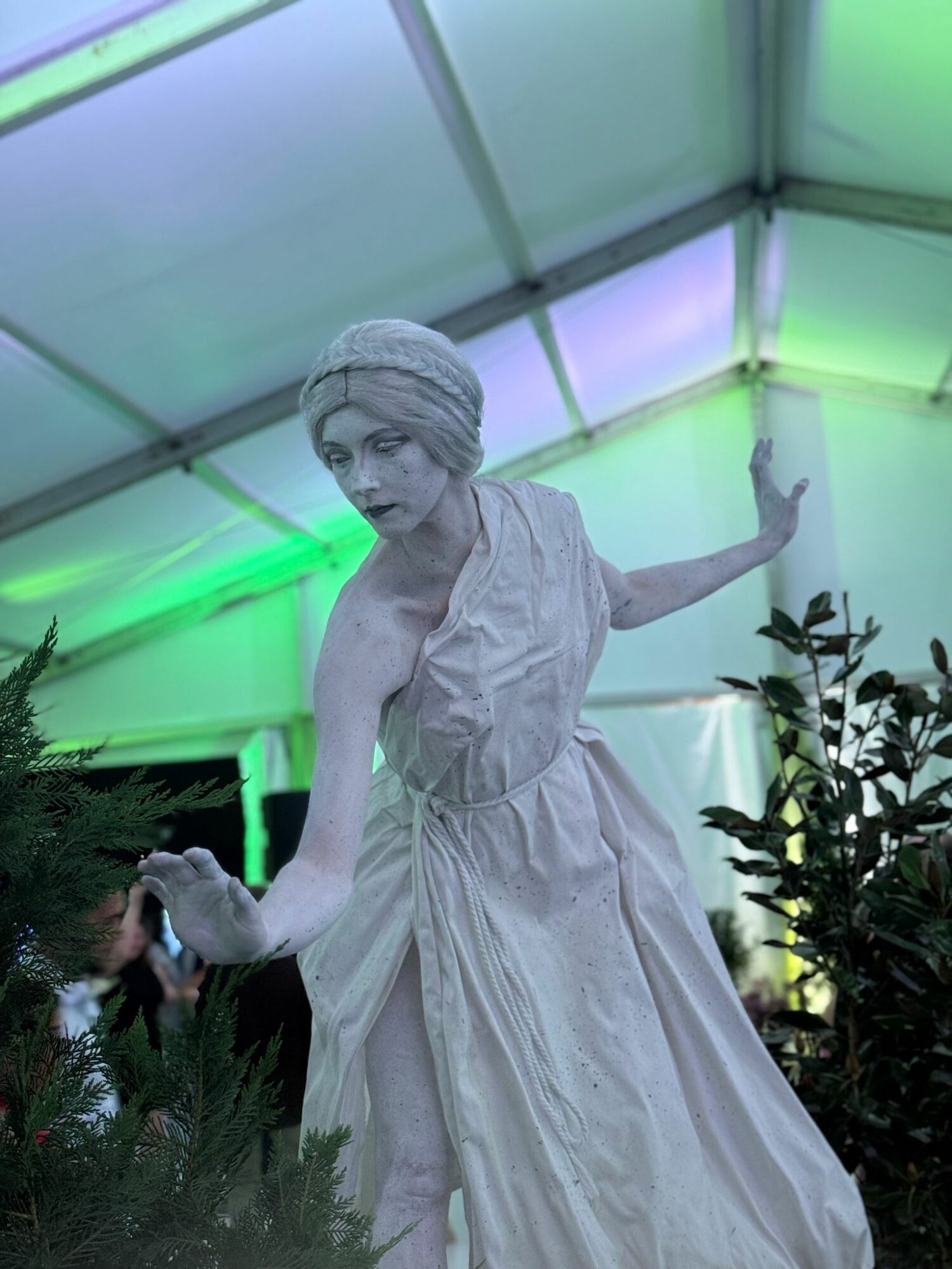 Statue of a woman in a flowing dress and headscarf, inside a tent with green and purple lighting, surrounded by plants.