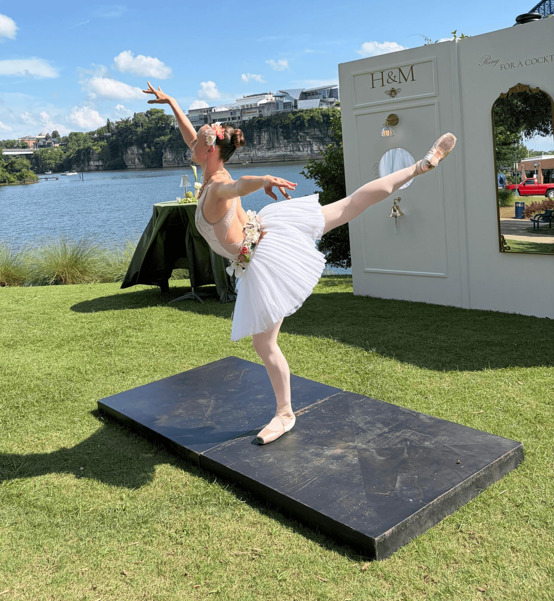 Ballet dancer in white tutu performs on a black platform outdoors near a river, with a white photo booth and blue sky.