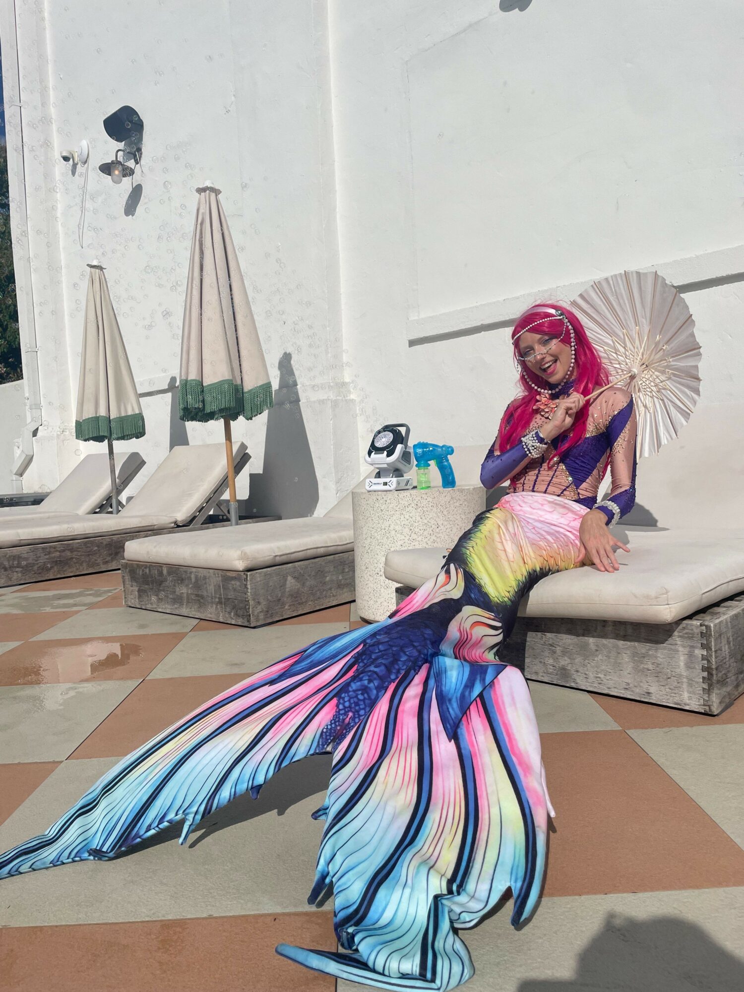 Person dressed as a mermaid with a colorful tail, pink hair, holding a fan, sitting on a bench outdoors.