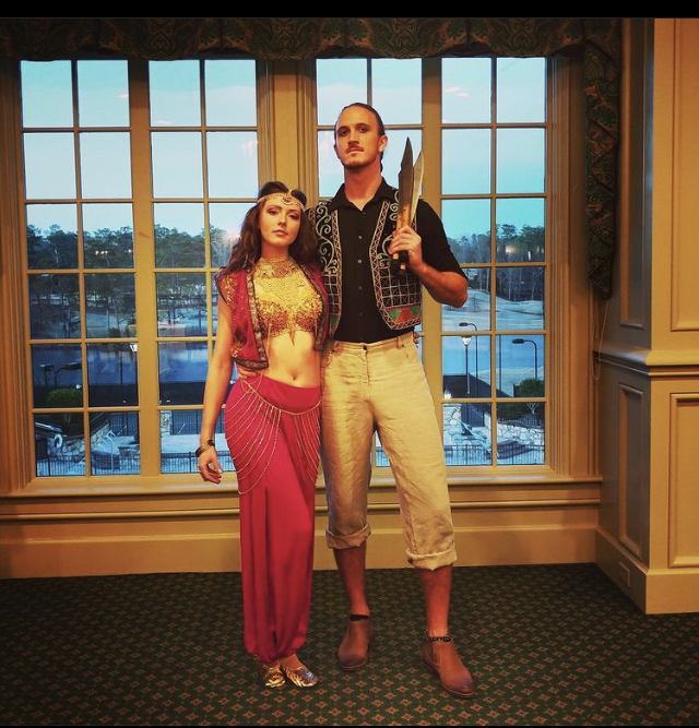 Two people stand in front of large windows with a view of a parking lot and trees, one holding a sword, both dressed in vintage-style clothing.