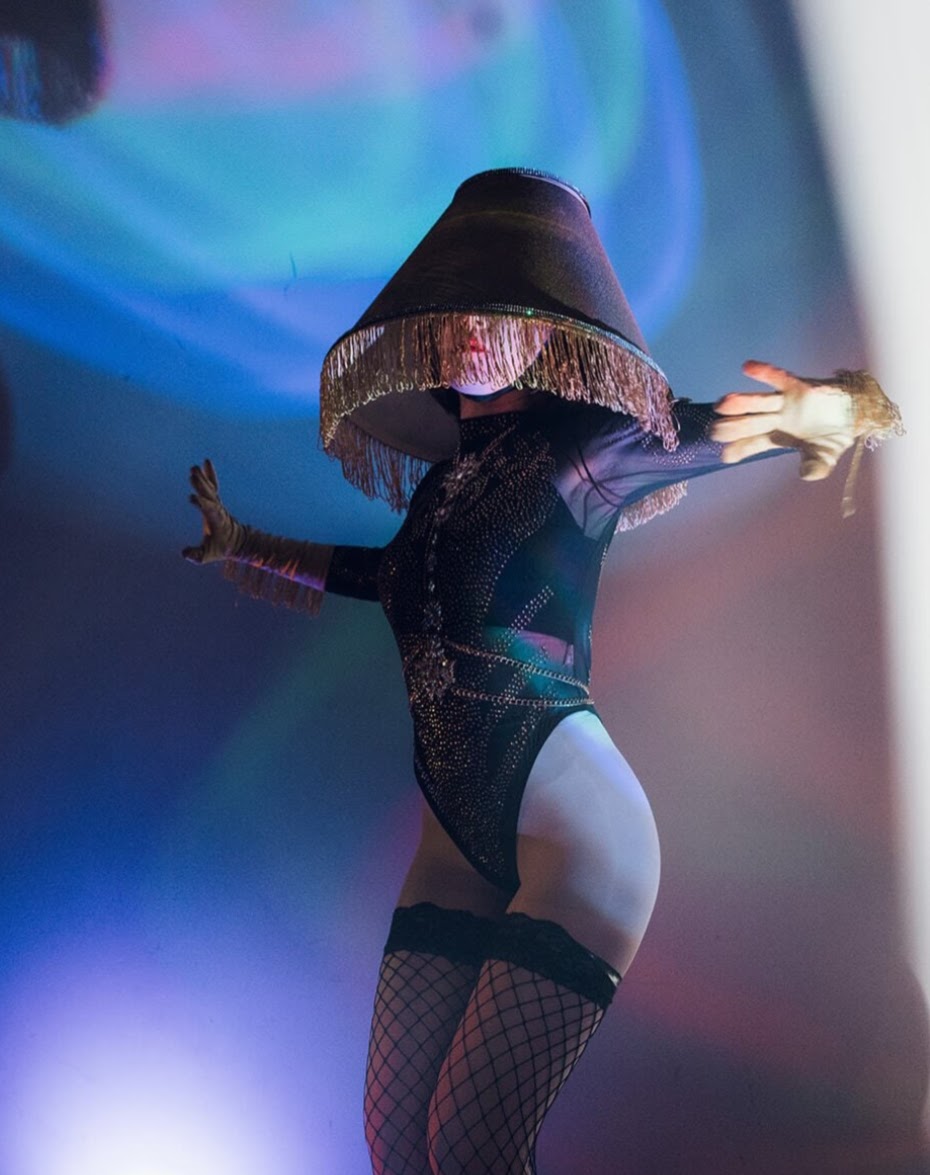 Person in costume with large hat, posing with arms outstretched, colorful background, fishnet stockings, and bodysuit.