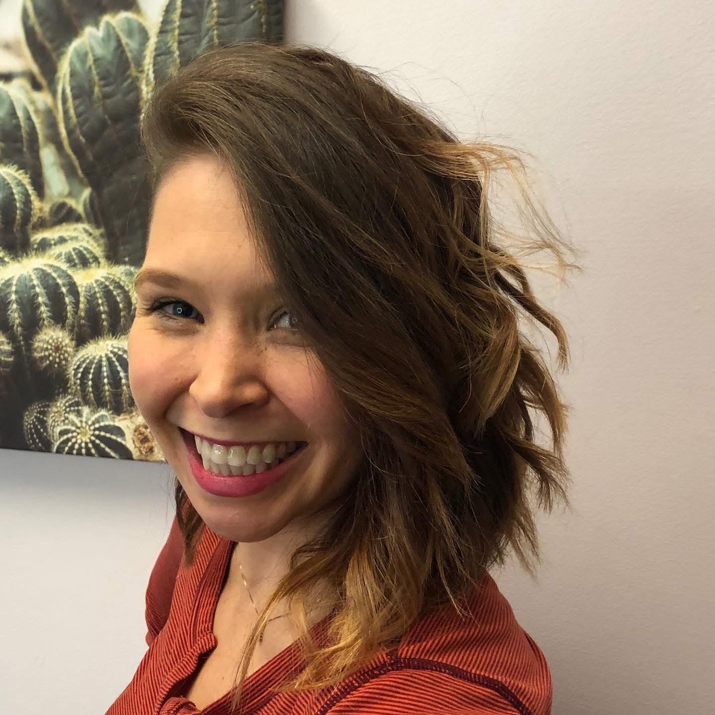Smiling woman with wavy hair wearing a red top, standing indoors near a cactus painting.
