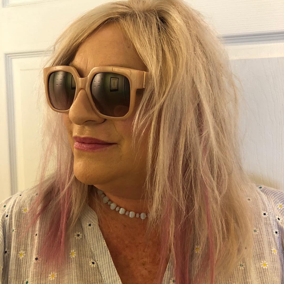 Woman with blonde hair, wearing large sunglasses, a pearl necklace, and a light-colored top with small patterns, smiling slightly.