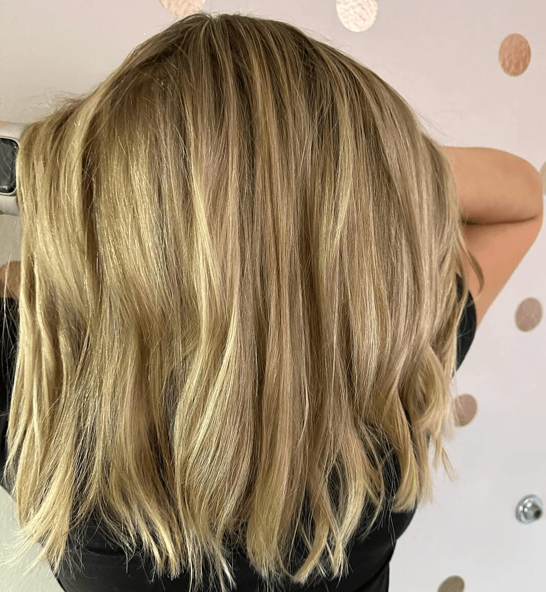 Back of a person's head with shoulder-length blonde hair, wavy and layered, against a light background with circular decorations.