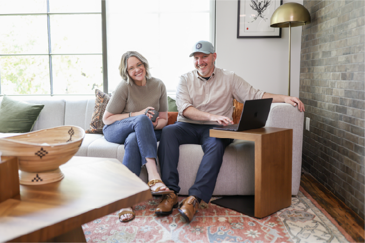 Two people sitting on a sofa, smiling, with a laptop on a small table, in a bright room with large windows.