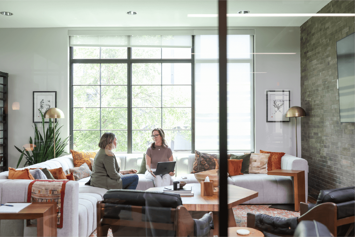 Two women sit on a sofa in a bright, modern living room with large windows, plants, and artwork.