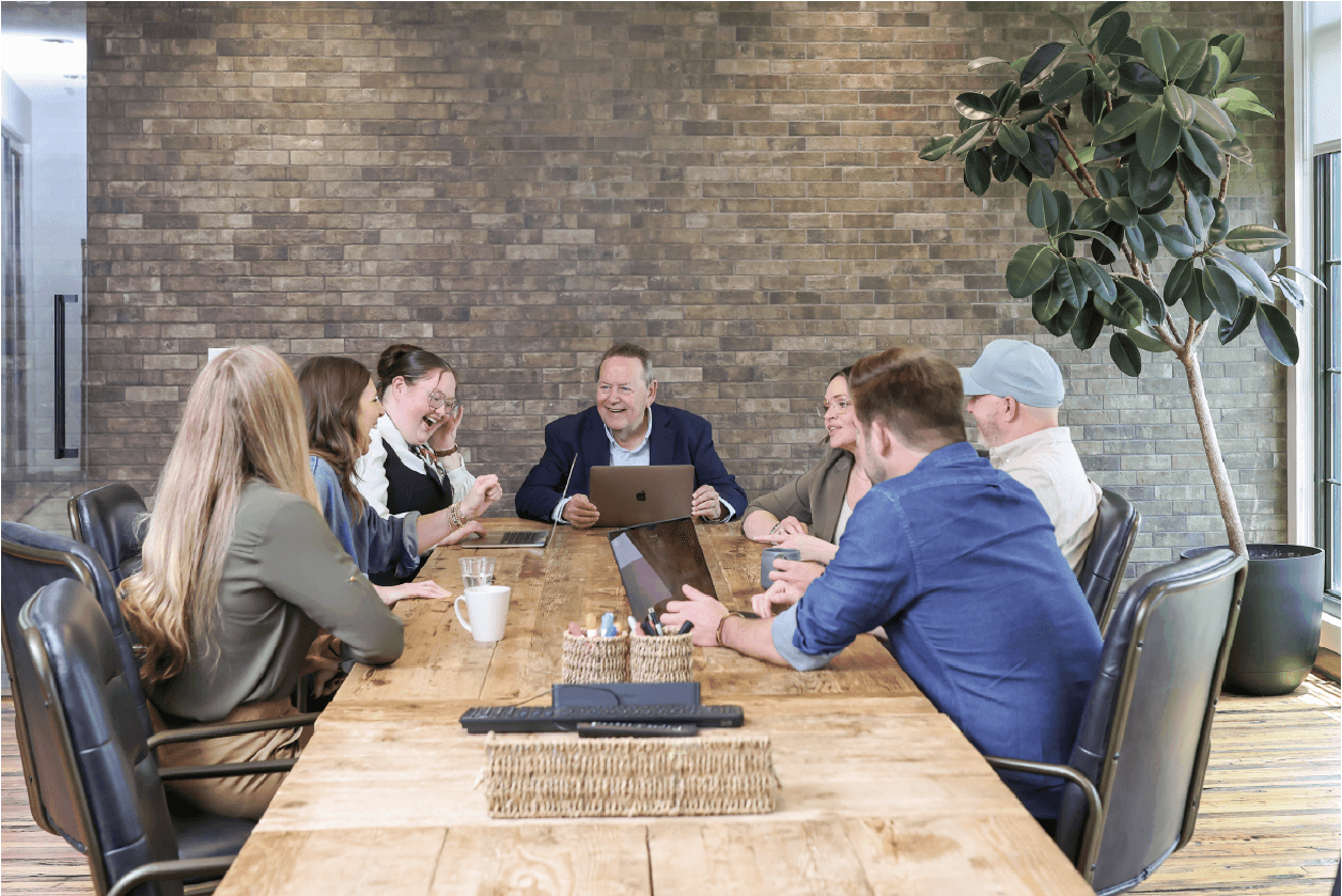 Group of seven people sitting around a wooden table in a modern office, engaged in discussion, with laptops and coffee cups present.
