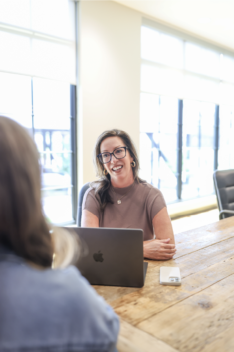 Woman with glasses smiling at a woman with brown hair, sitting at a wooden table with a laptop and phone, in a bright room.