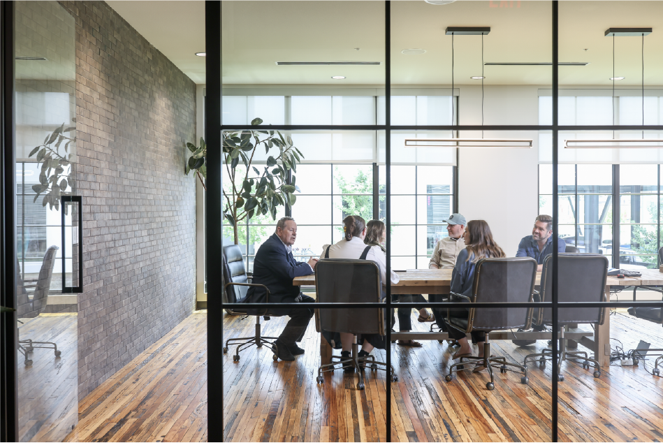 Group of people sitting around a table in a modern conference room with large windows and wooden floors.