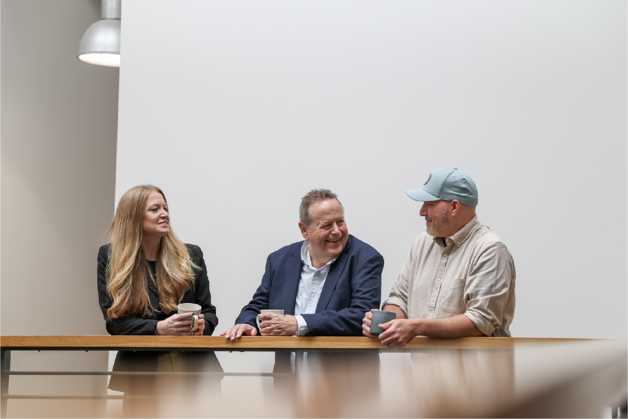 Three people sitting at a table, smiling and talking, with a plain white background.