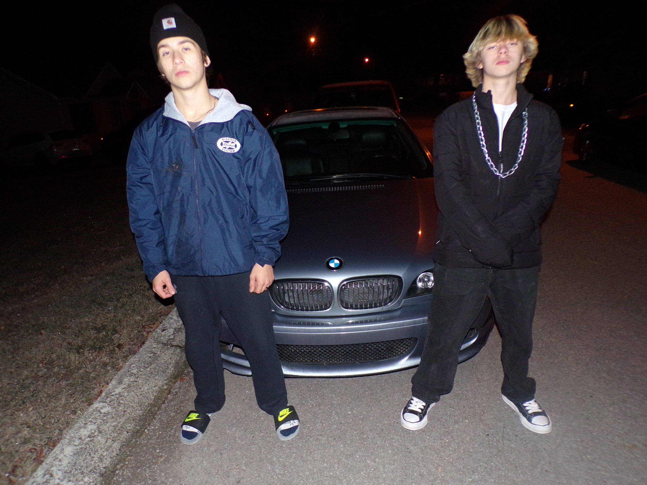 Two young people stand in front of a car at night, one wearing a blue jacket and beanie, the other in black with a necklace.